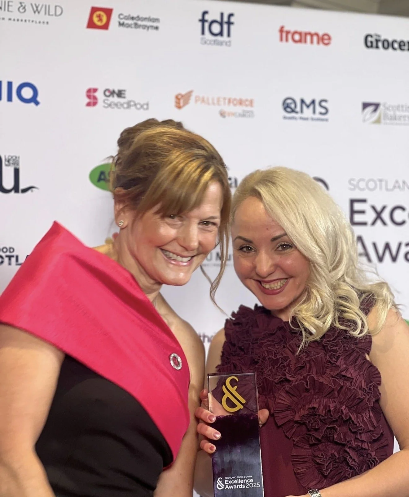 Two women at an awards event, smiling and holding a trophy, with a backdrop displaying various sponsor logos and text such as 'SCOTLAND FOOD & DRINK Excellence Awards 2025'.