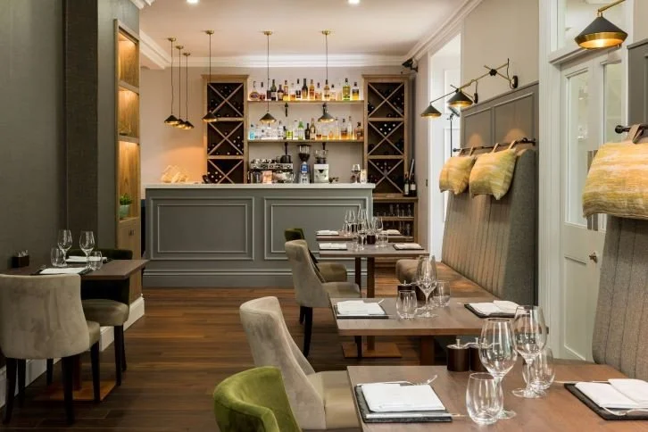 Elegant restaurant with wooden floors, beige and green upholstered chairs, a bar with wine racks, and tables set with glasses, plates, and napkins.