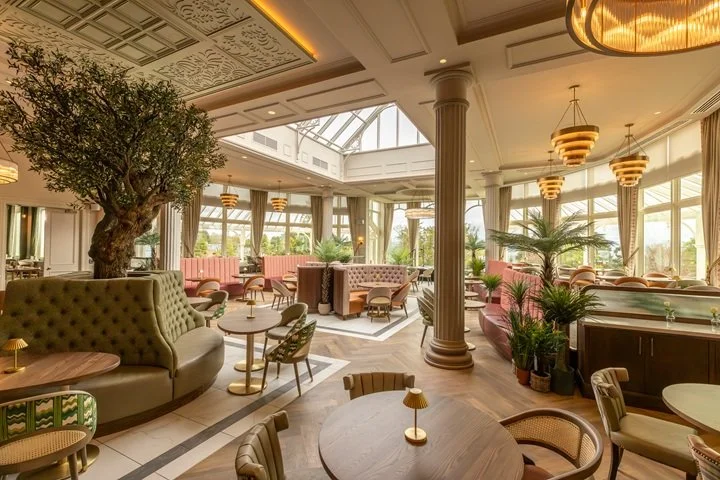 Elegant lounge with large windows, classical columns, potted plants, round wooden tables, upholstered chairs, and soft lighting.