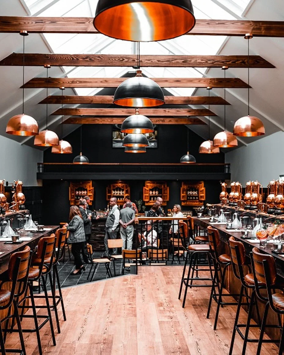 Interior of a modern restaurant or bar with wooden beams on a high ceiling, hanging copper and black pendant lights, a bar area with shelves of glasses, and several groups of people socializing.