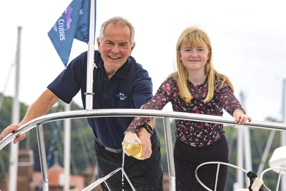 An older man and a young girl smiling on a boat, with the man holding the railing and the girl holding a glass of drink.