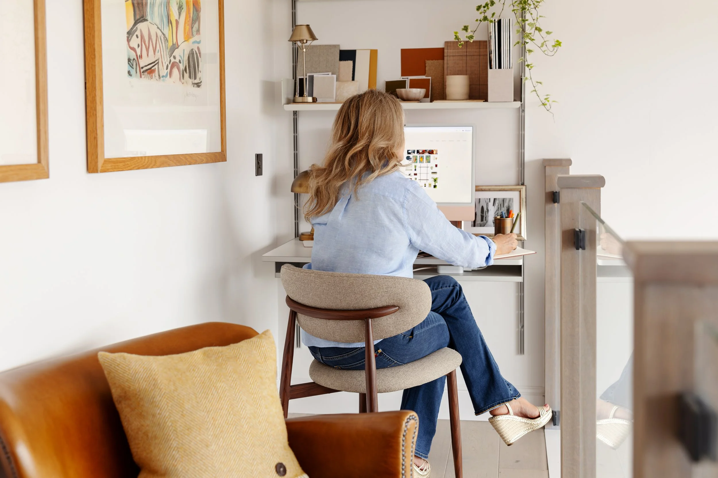 A woman with wavy blonde hair, wearing a light blue shirt and dark jeans, sitting at a small desk in a bright room. She is facing away from the camera and working on a computer with a white background, which displays images and videos. The room has artistic decor, a beige upholstered chair, a leather armchair with a yellow pillow, and shelves with books, plants, and framed artwork.