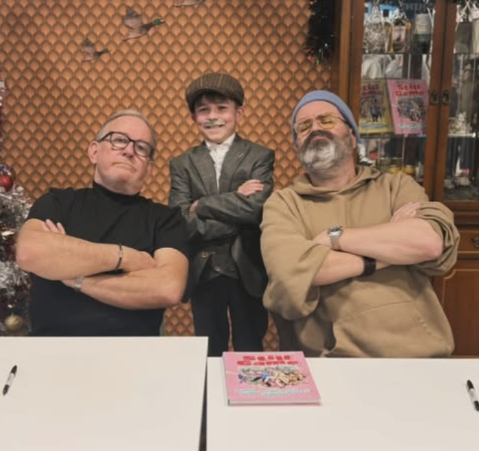 Three men posing with their arms crossed, two of them with painted faces, standing behind a white table with a pink book on it, in front of a wooden wall with decorations and bookshelves.
