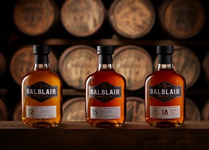 Three bottles of Balblair Scotch whisky on a wooden surface with barrels in the background, labeled with different ages: 12, 15, and 18 years.