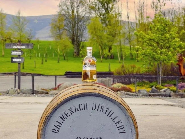 A bottle of Caorunn gin atop a whiskey barrel outdoors in a green park with trees and hills in the background.
