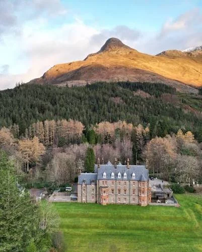 A large, historic castle-like building with a gray roof is situated in front of a forest and a mountain range in the background.