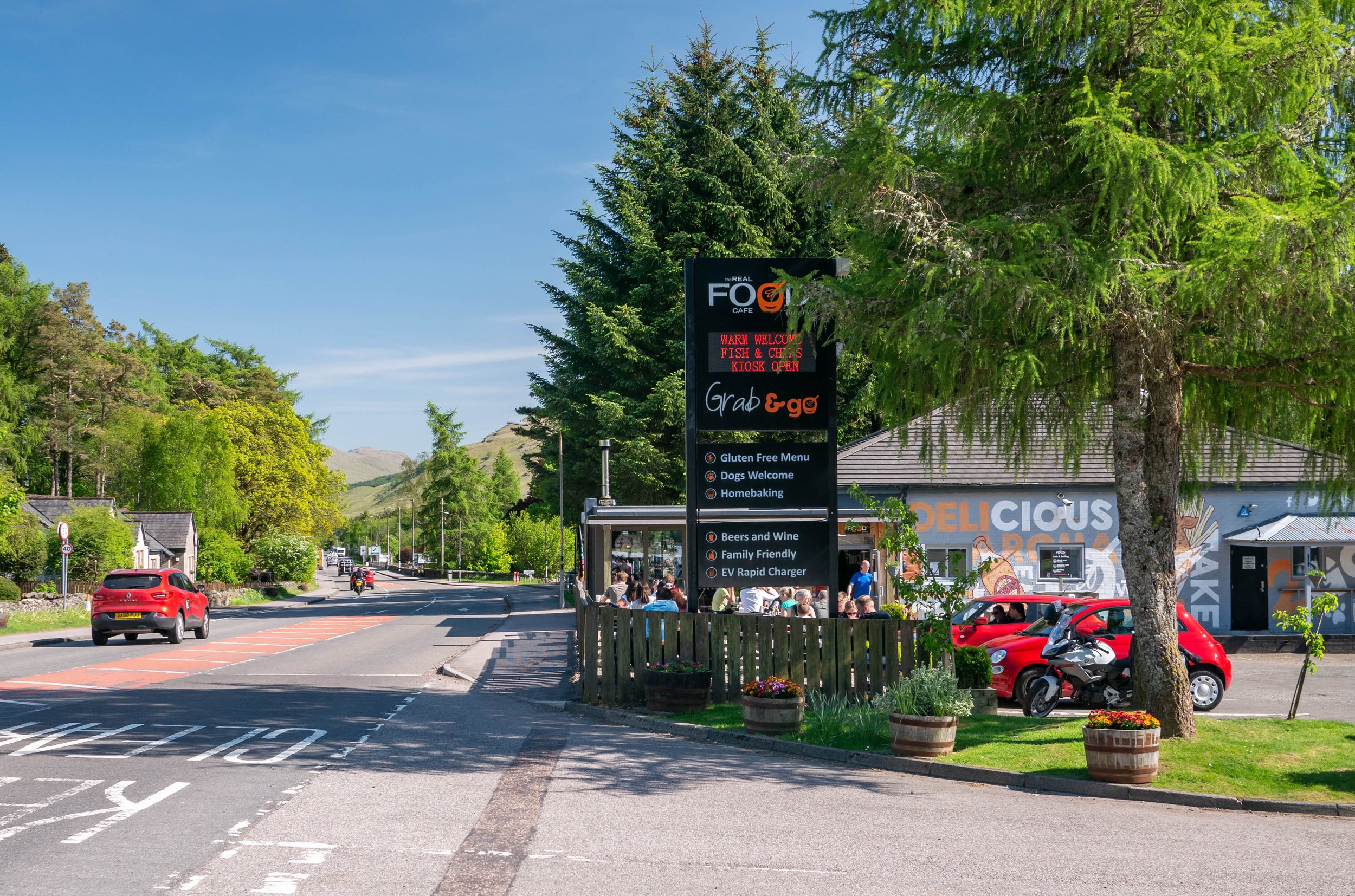 A street scene near a cafe with a sign advertising food and drinks, surrounded by trees and parked cars under a clear blue sky.