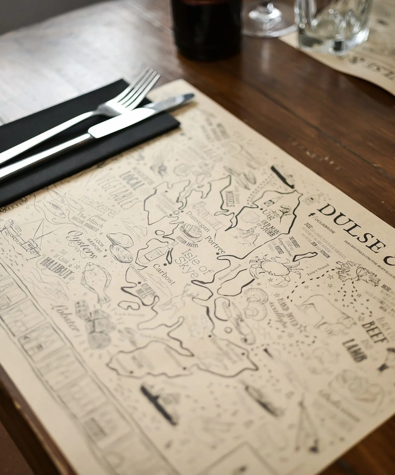 A detailed black and white illustrated map of Dumfriesshire in Scotland on a wooden table, with a set of silverware placed on a black napkin nearby.
