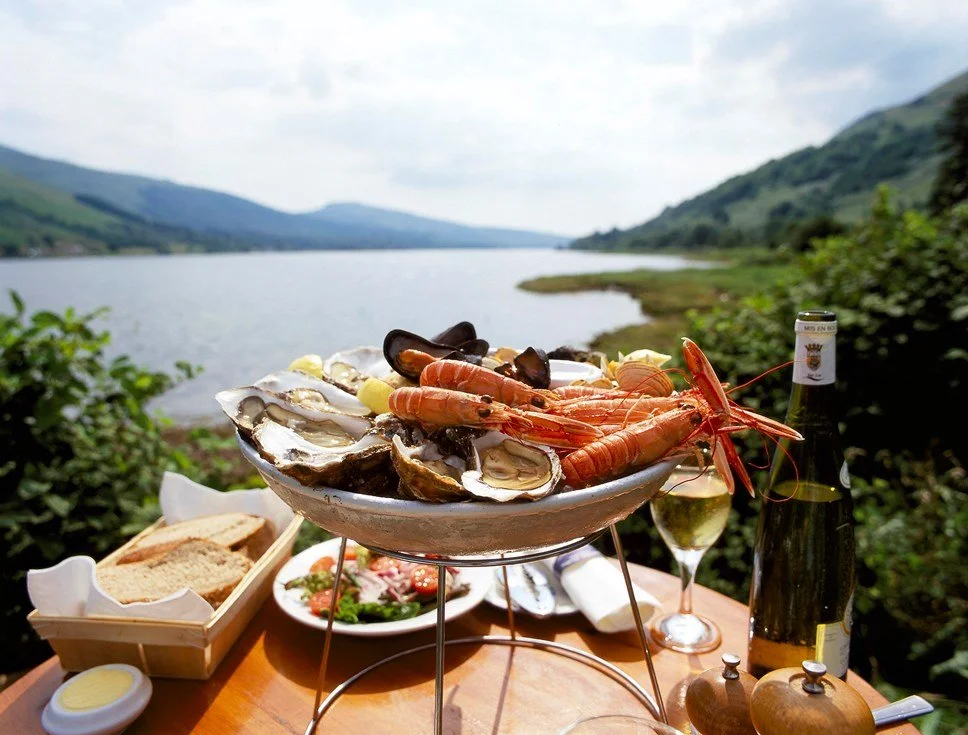 Seafood platter with oysters and lobster on an outdoor table overlooking a lake or river with green hills in the background.