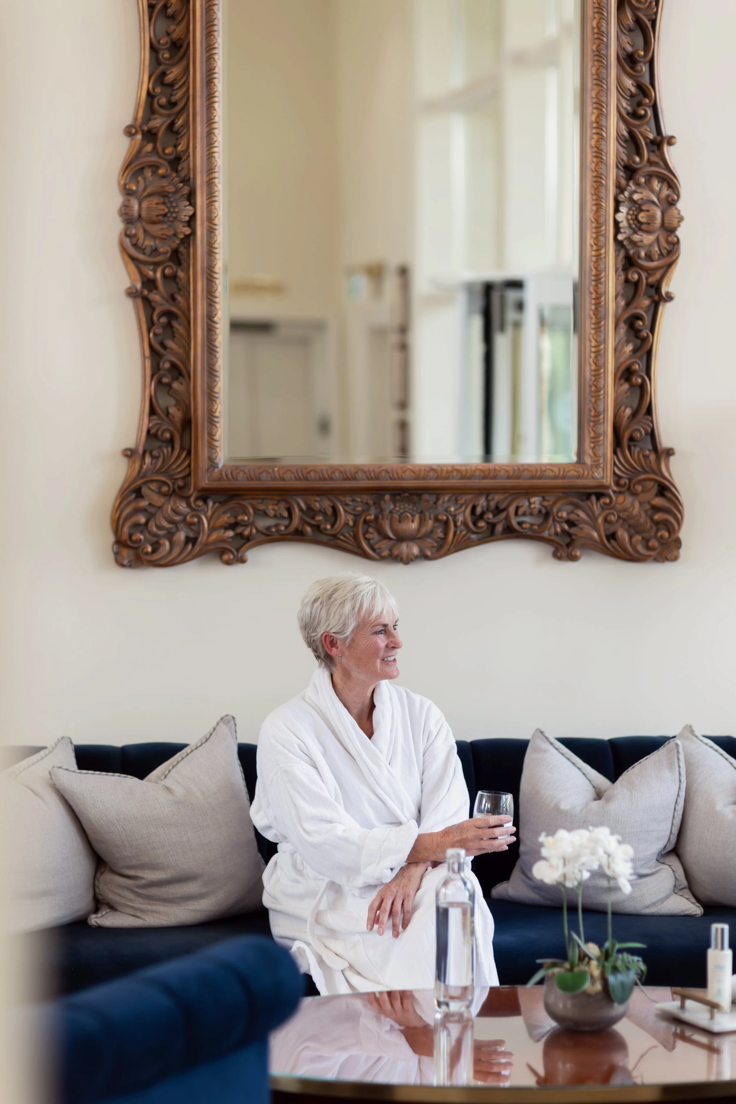 A woman with short gray hair wearing a white bathrobe sitting on a beige couch with beige pillows, holding a glass of water, in a living room with a large ornate mirror on the wall and a dining table with a flower arrangement and water bottles in the foreground.