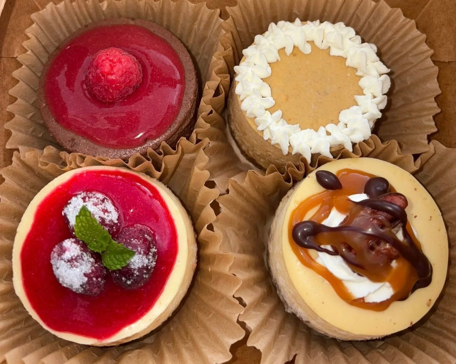 Assorted handcrafted mini cheesecakes in a bakery box, featuring raspberry chocolate, pumpkin, cranberry, and caramel pecan flavors from Lil C’s Mini Cheesecakes