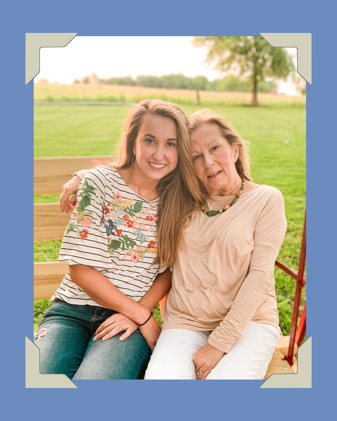 Two women sitting close together on a wooden bench outdoors, smiling, with green grass and trees in the background.