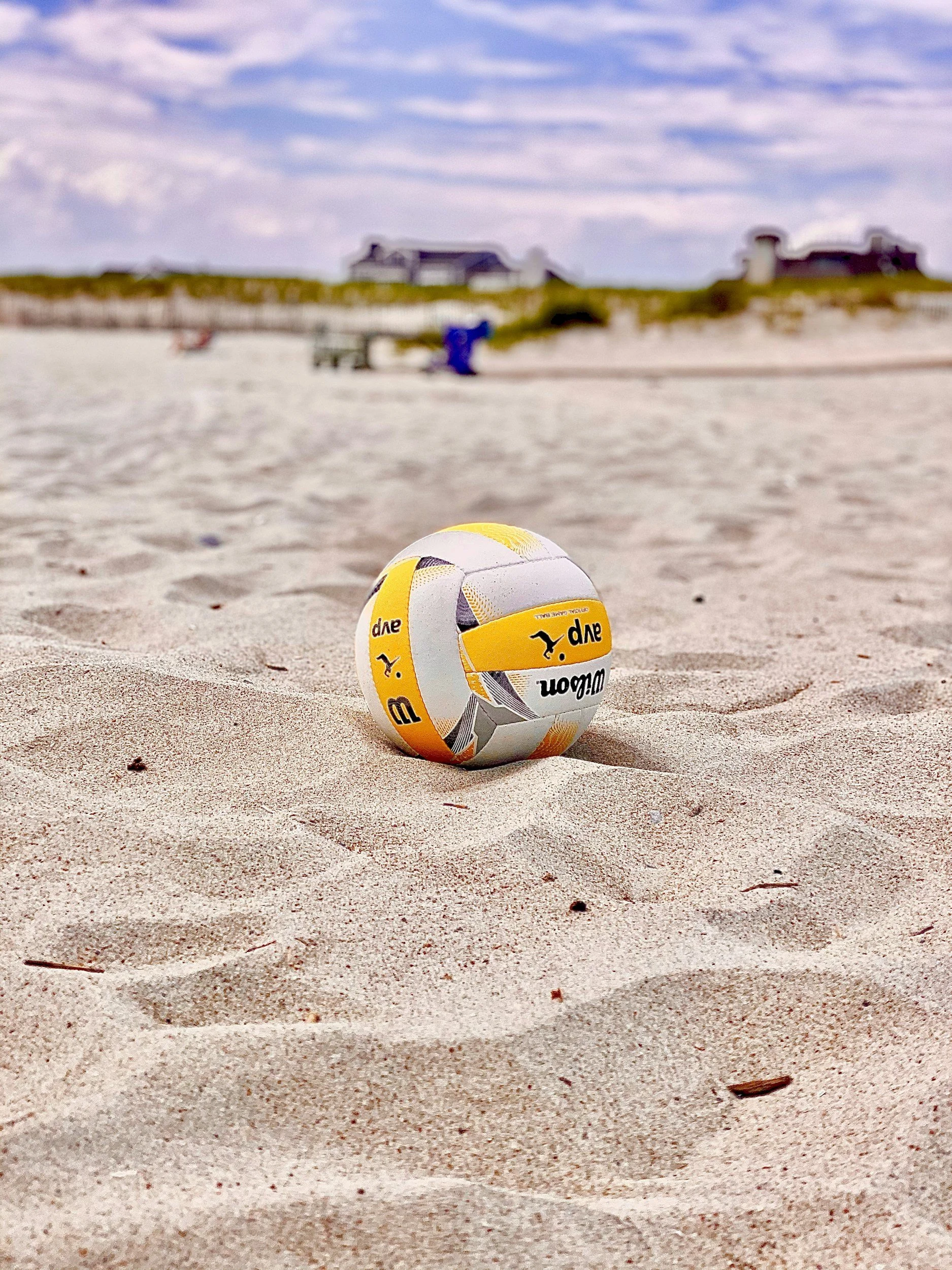 A yellow and white volleyball resting on the sandy beach with footprints and small debris, with houses on a hill in the background under a partly cloudy sky.