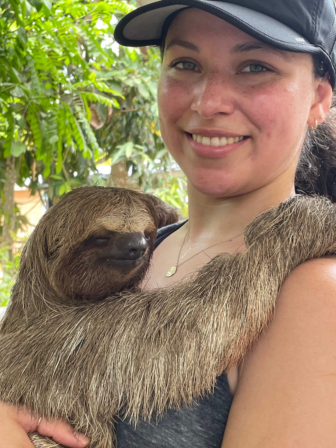 A woman holding a sloth on her arm outdoors with green foliage in the background.