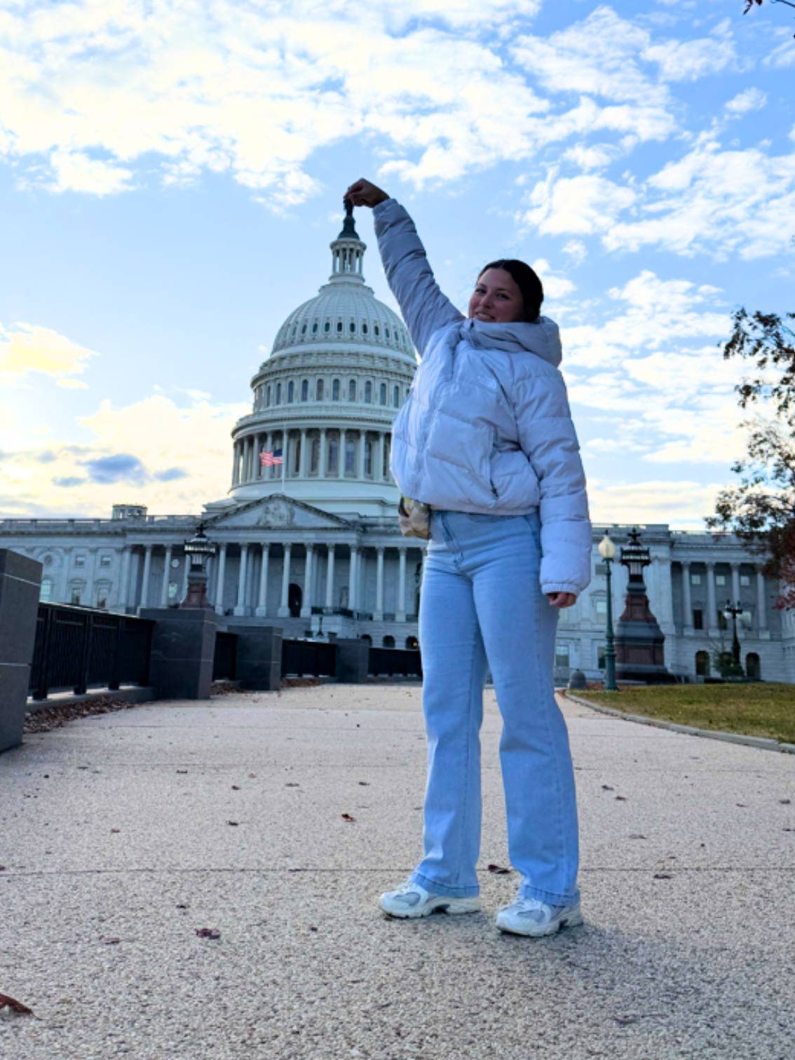 Woman in white jacket and blue jeans standing in front of the U.S. Capitol building, holding an object in the air, with a partly cloudy sky.