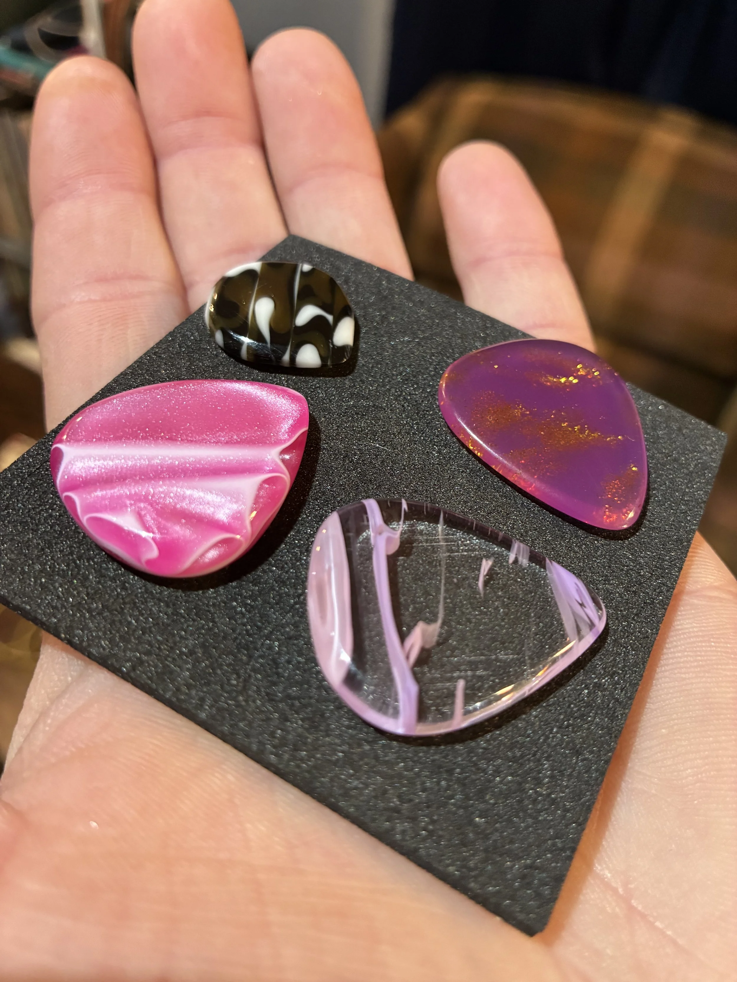 Close-up of a person's hand holding a black foam display with five colorful guitar picks made of resin, in pink, purple, mottled black and white, and marble-like designs.