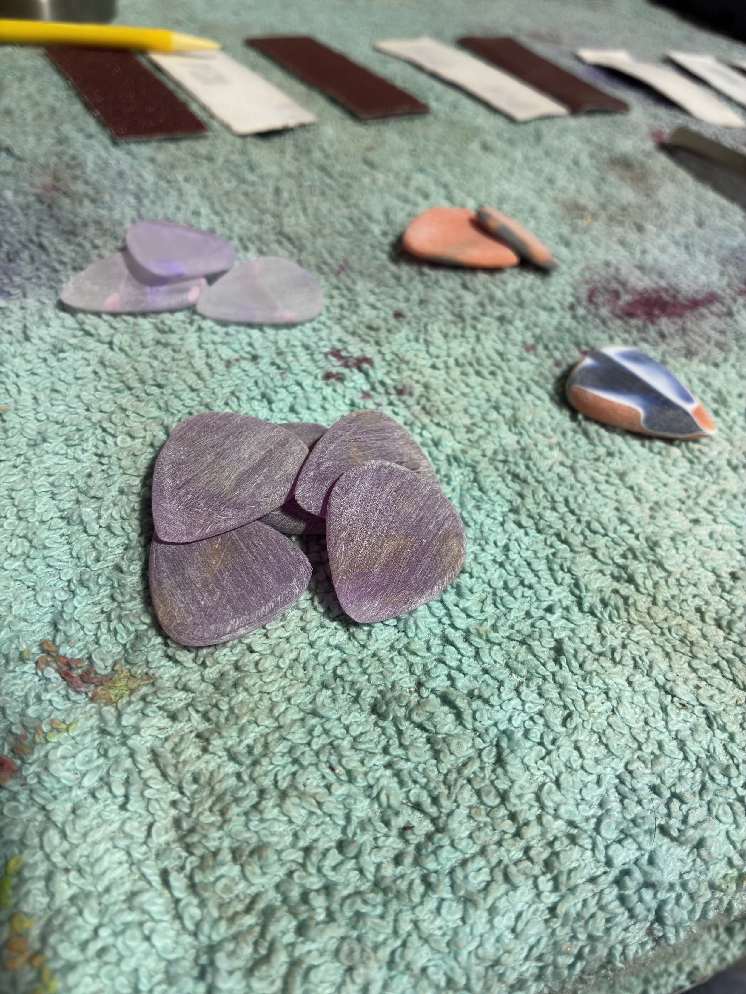 Various guitar picks in different colors, shapes, and sizes on a textured green surface, with some rectangular objects in the background.