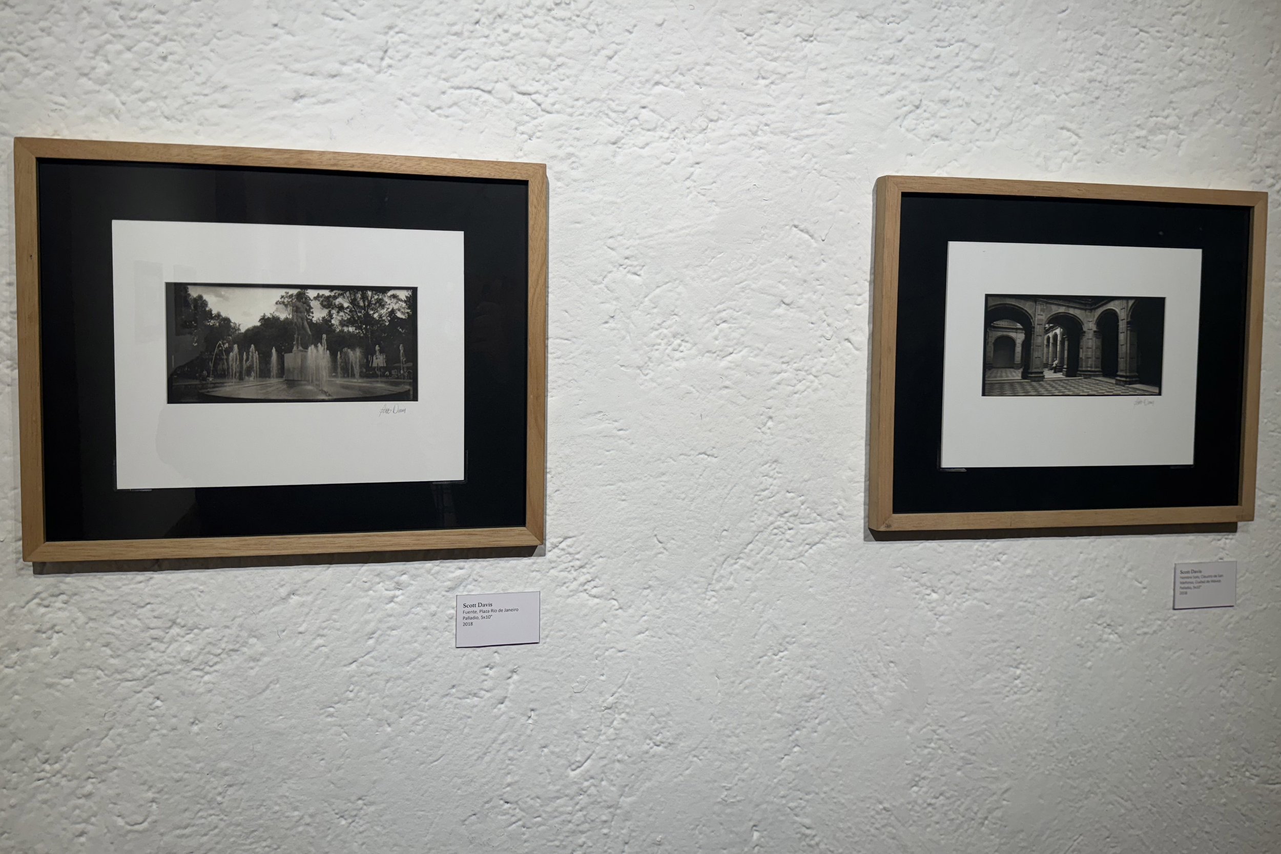 Two framed black and white photographs hung on a textured white wall. The left photo depicts a fountain with trees in the background, and the right photo shows an architectural corridor with arches and columns.