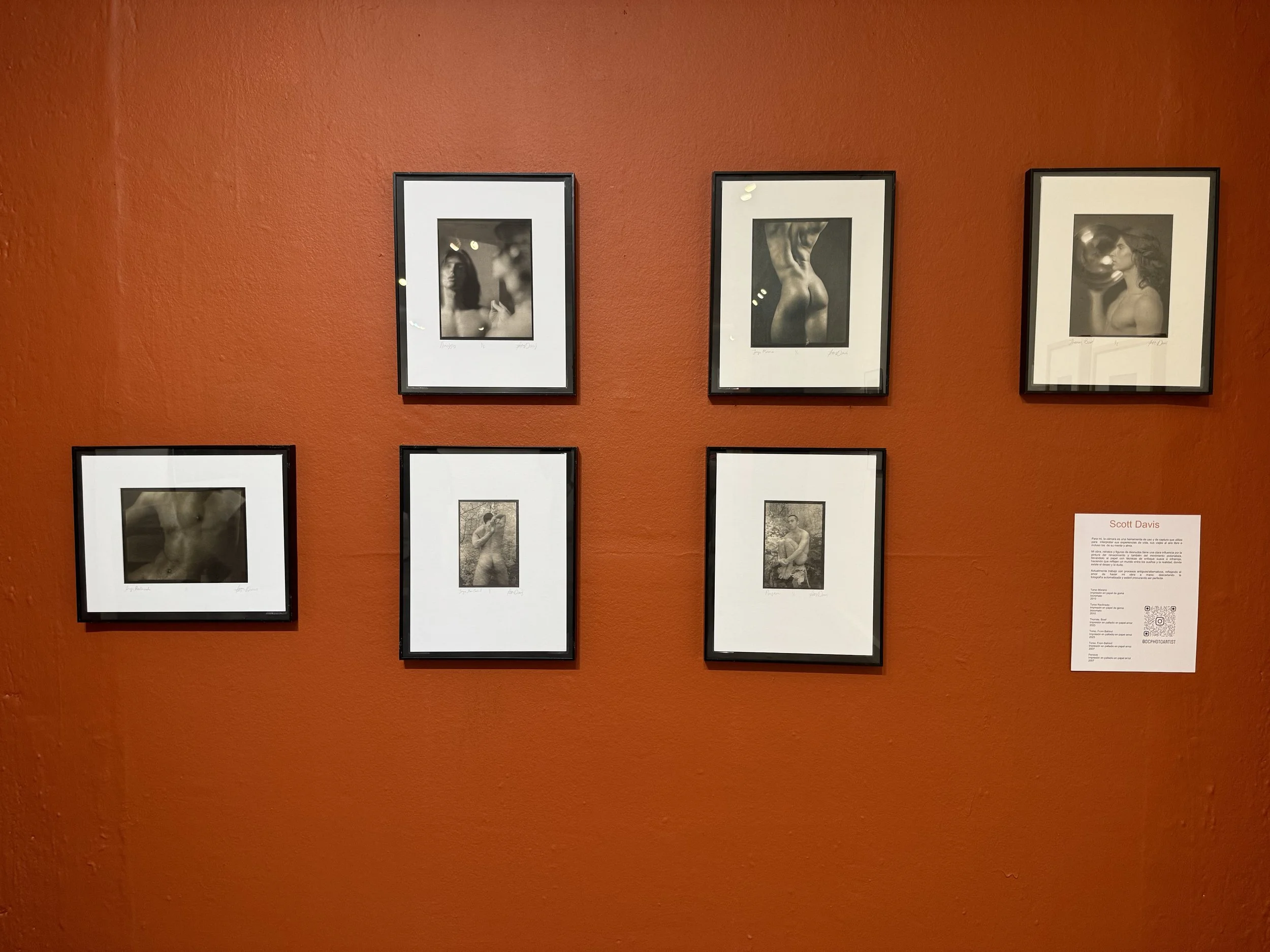 Art gallery wall with framed black-and-white photographs of nude and semi-nude models, including a woman holding a mask and a person with a visible back, labeled with the artist's name Scott Davis.