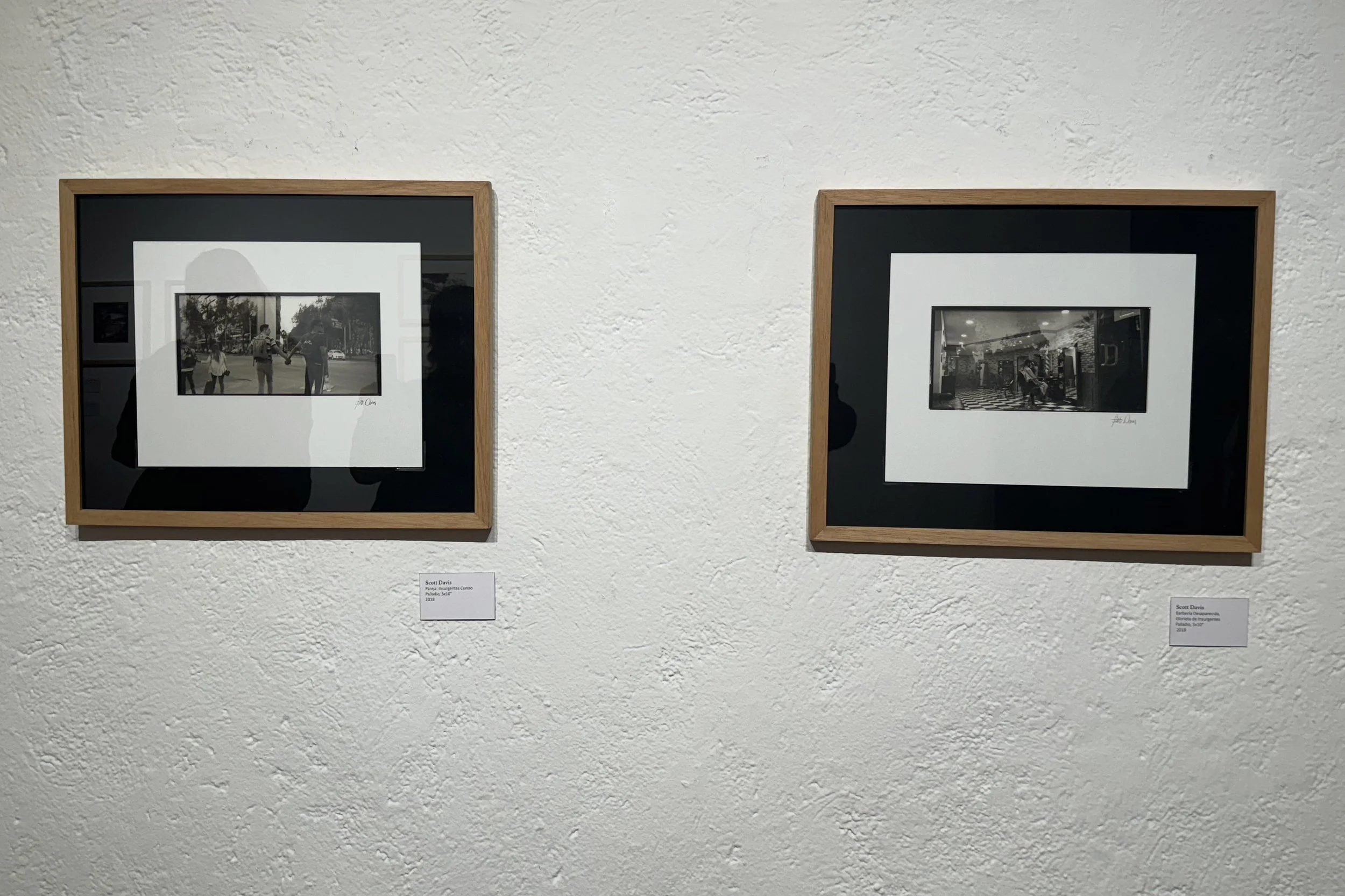 Two framed black-and-white photographs hanging on a white textured gallery wall, each accompanied by a small label below.