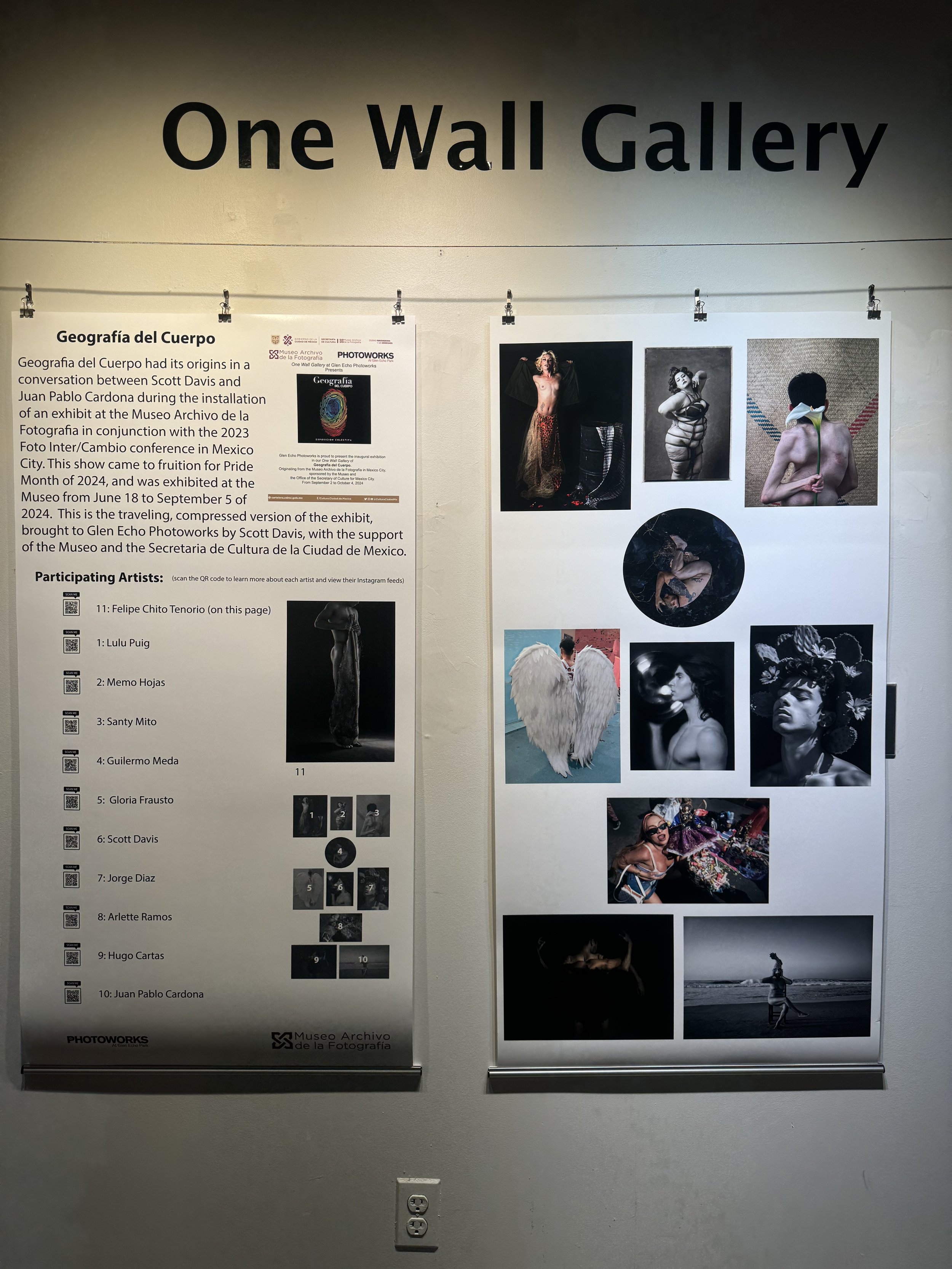 Exhibition display board titled 'One Wall Gallery' presenting photographs by various artists. The left section contains an informational paragraph about the exhibit, mentioning Scott Davis and the Museo Archivo de la Fotografía. The list of participating artists includes Felipe Chito Tenorio, Lulu Puig, Memo Hojas, Santy Mito, Guillermo Meda, Gloria Frausto, Jorge Diaz, Arlette Ramos, Hugo Cartas, and Juan Pablo Cardona. The right section showcases photographic artworks featuring diverse subjects, including a topless woman with elaborate makeup, a woman with angel wings, a person with a flower mask, and other artistic portraits.