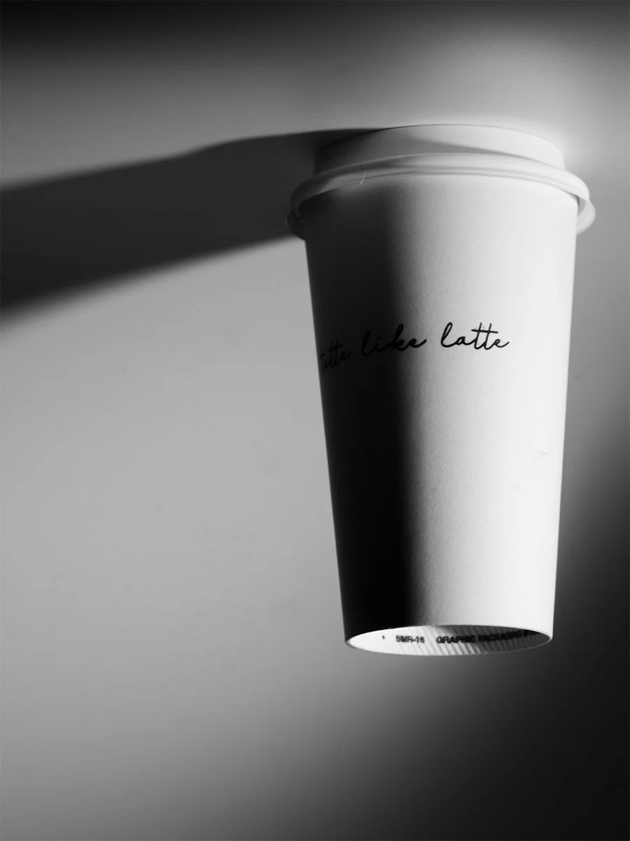 A white disposable coffee cup with a lid, suspended in the air, casting a shadow on the wall behind it. The cup has black handwritten-style text that says "just like latte".