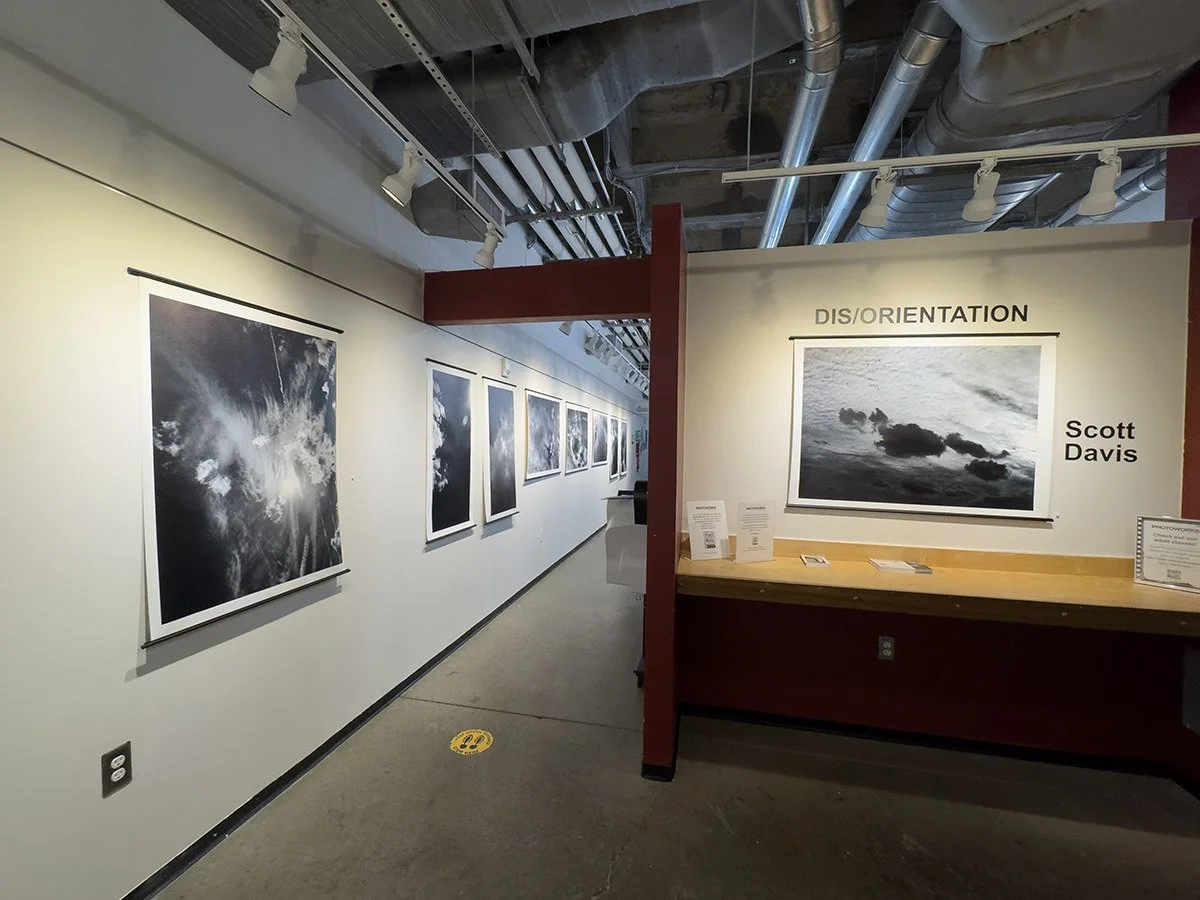Black and white photography exhibit with various cloud and sky images on white walls, featuring a section titled 'DIS/ORIENTATION' by Scott Davis.