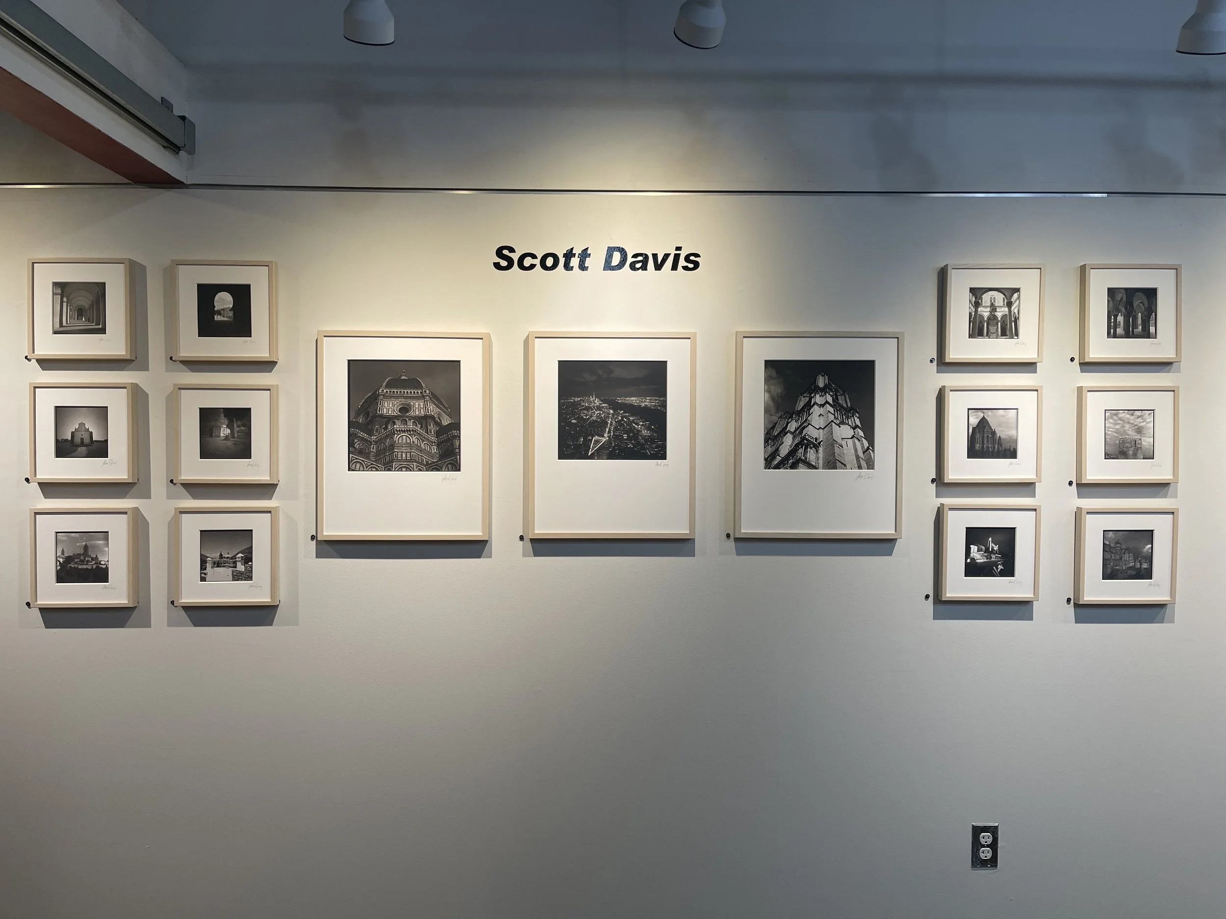 Gallery wall titled 'Scott Davis' featuring black and white photographs in frames, displayed on a white wall with overhead lighting.