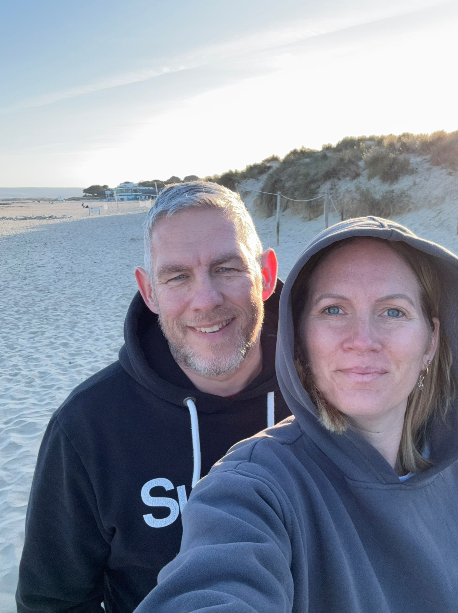 A man and a woman taking a selfie on a beach during sunset. The man has gray hair and a beard, wearing a black hoodie, and the woman has blue eyes, brown hair, and is wearing a gray hoodie.