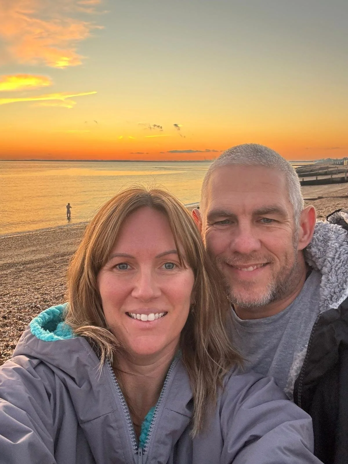 A smiling couple taking a selfie on a beach at sunset with the ocean and a person standing in the water in the background.