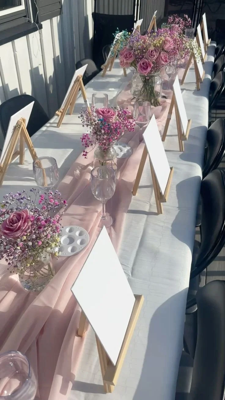 A long banquet table decorated with pink and purple flowers in glass vases, wooden framed blank whiteboards, transparent glasses, and a pink satin table runner, set in a sunlit room for a celebration or event.
