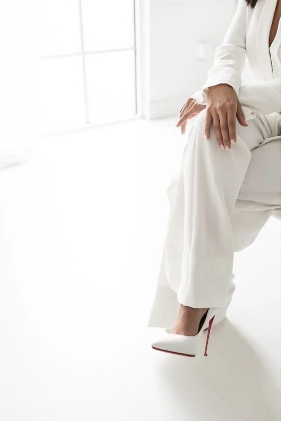 A person dressed in a white suit with high heels sitting in a bright room.