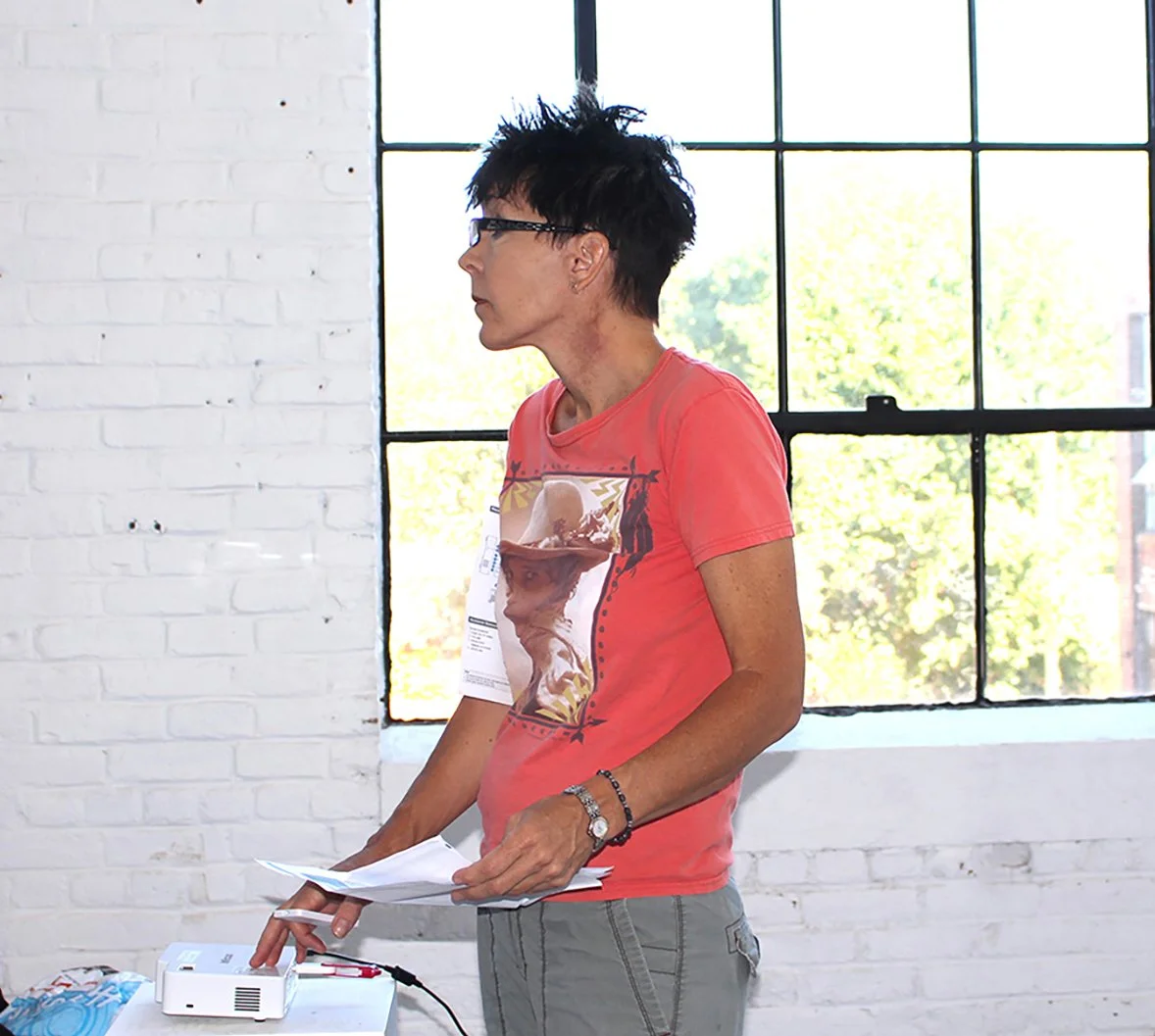 Woman with short black hair and glasses wearing a red t-shirt and gray pants, holding papers, standing in front of a large window with a view of trees outside.