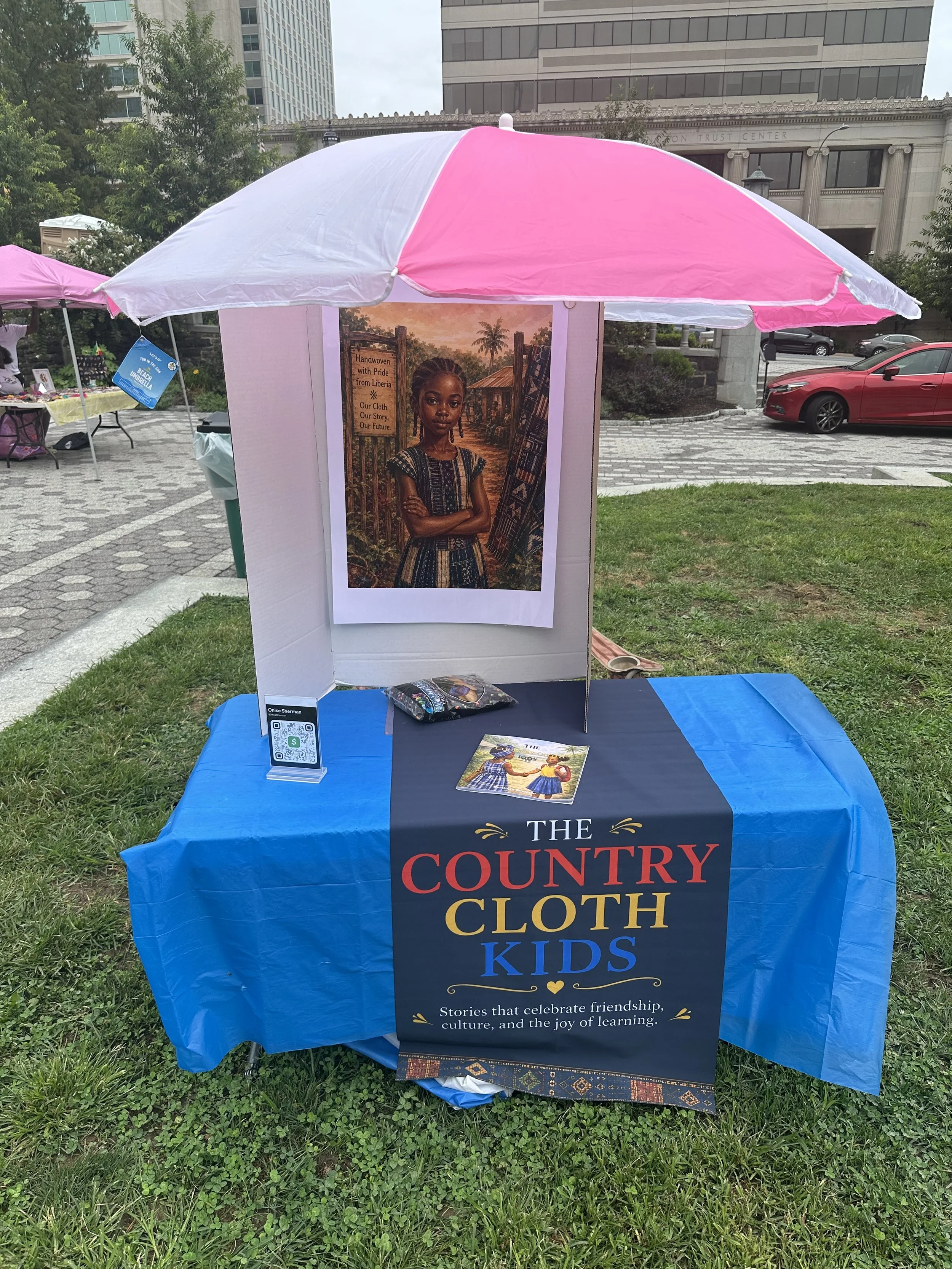 A booth at an outdoor event with a blue tablecloth that has the words "The Country Cloth Kids" printed on it. On the table is a small stack of books and a QR code stand. Behind the table is a large poster of a young girl with her arms crossed, standi