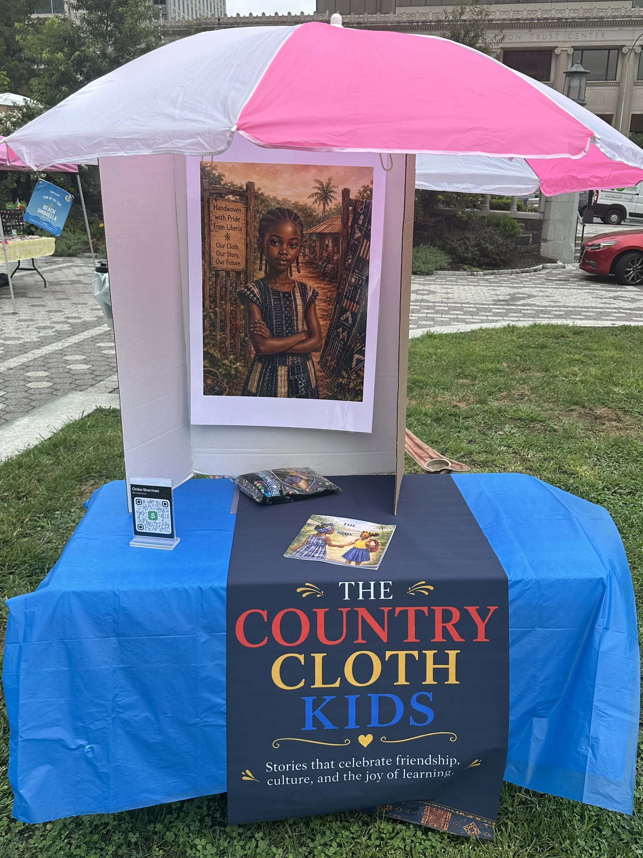 Display booth with a large poster of a young girl in traditional African clothing, a table with a black cloth featuring the words "The Country of Cloth Kids," and an umbrella overhead. The booth promotes stories that celebrate friendship, culture, an
