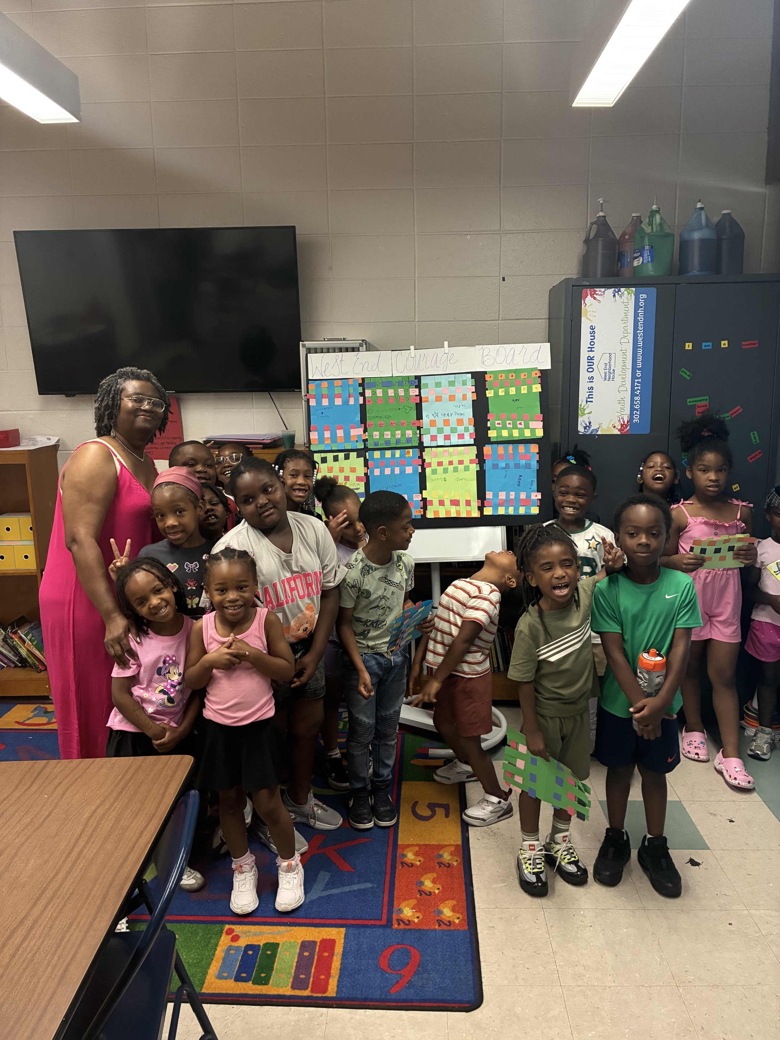A group of children and a woman standing in a classroom in front of a colorful board with sticky notes. The children are smiling and holding notes or making peace signs. A large television is mounted on the wall and lockers are visible in the backgro