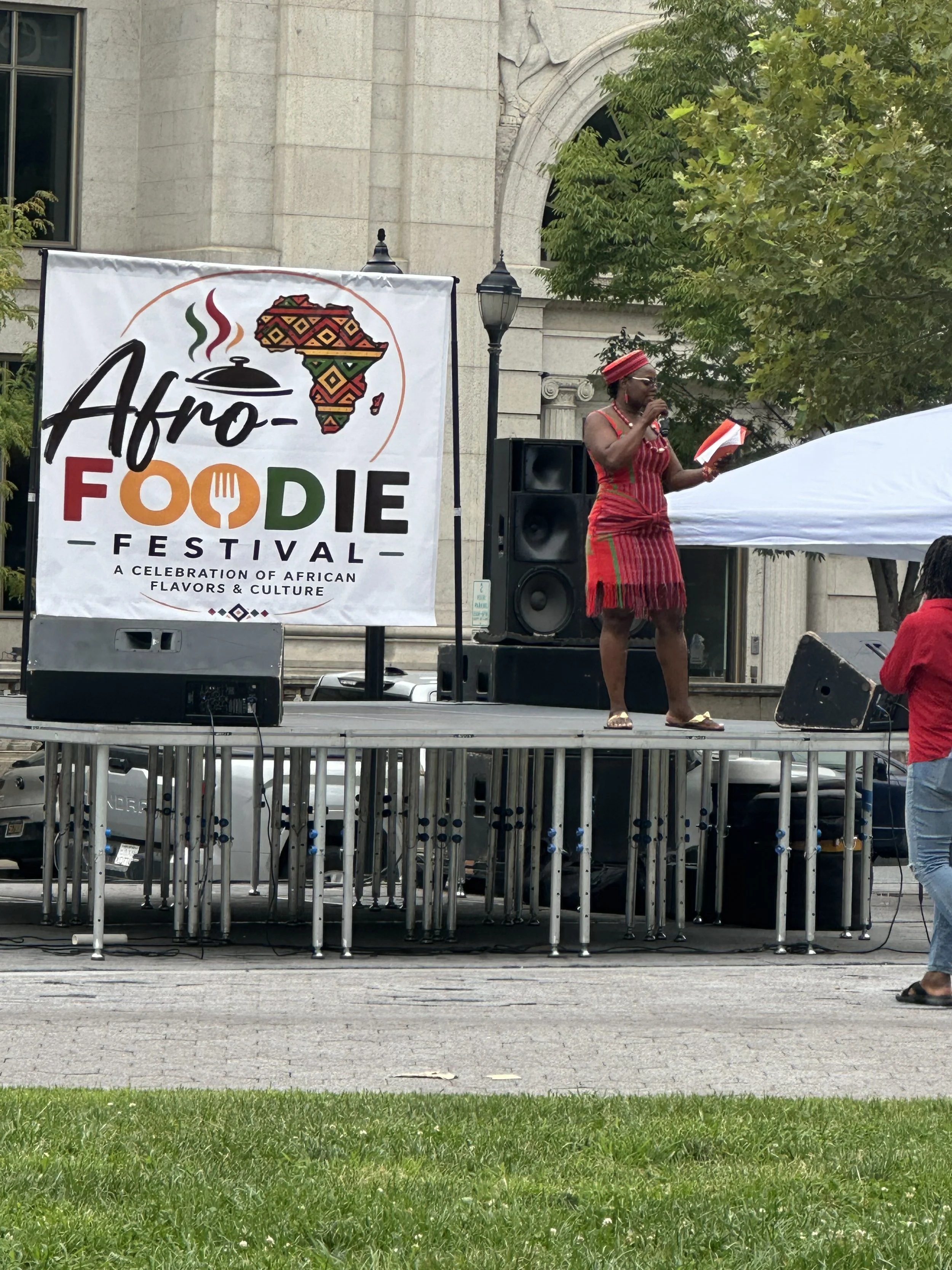 A woman dressed in traditional African clothing standing on an outdoor stage, speaking into a microphone, during the Afro-Foodie Festival, which celebrates African flavors and culture. The stage has a banner with the event name, a stylized map of Afr