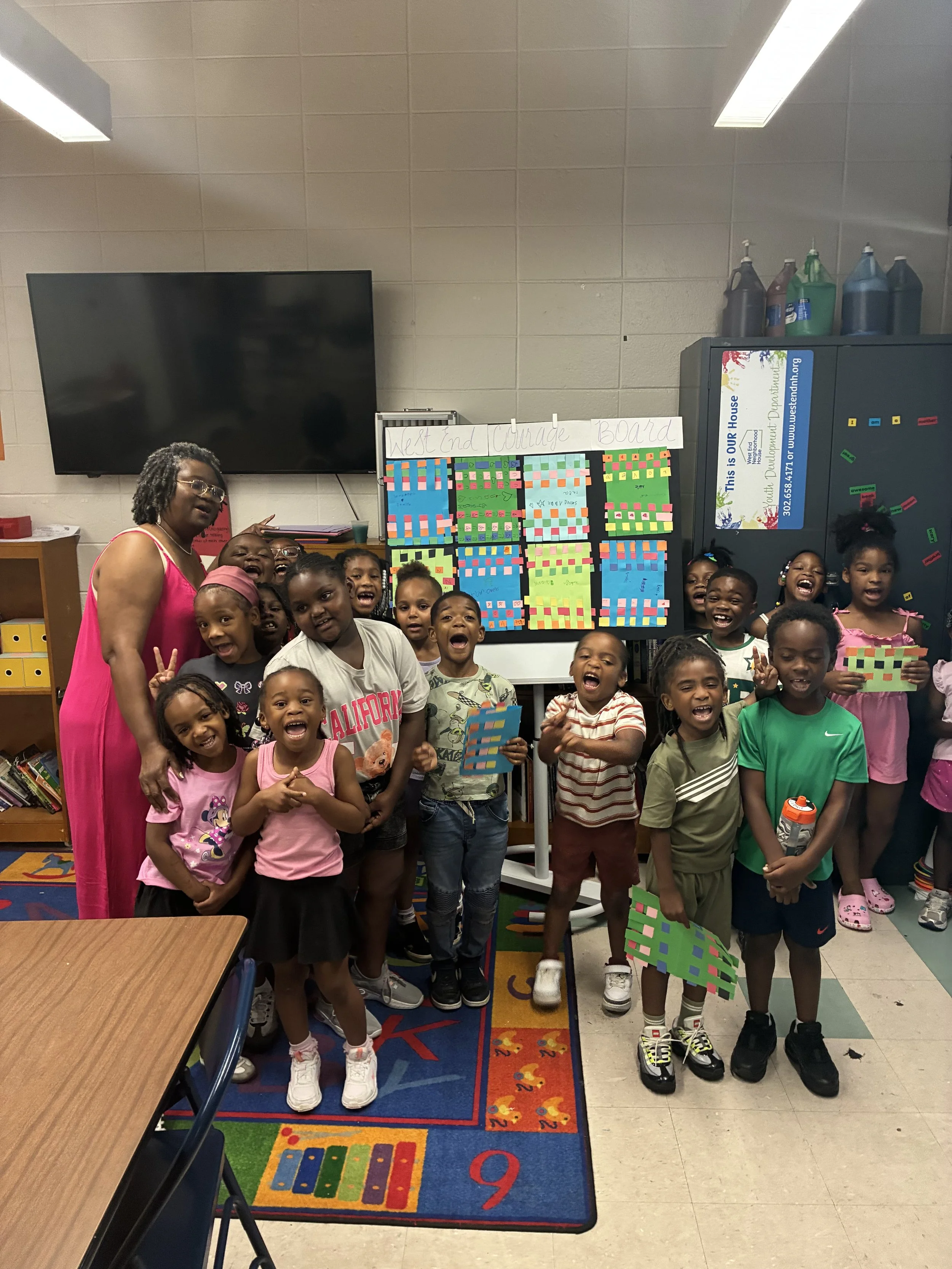 A group of young children with a woman in a classroom, posing in front of a whiteboard with colorful charts and a locker. The children are smiling and holding project papers, with a vibrant educational rug on the floor.