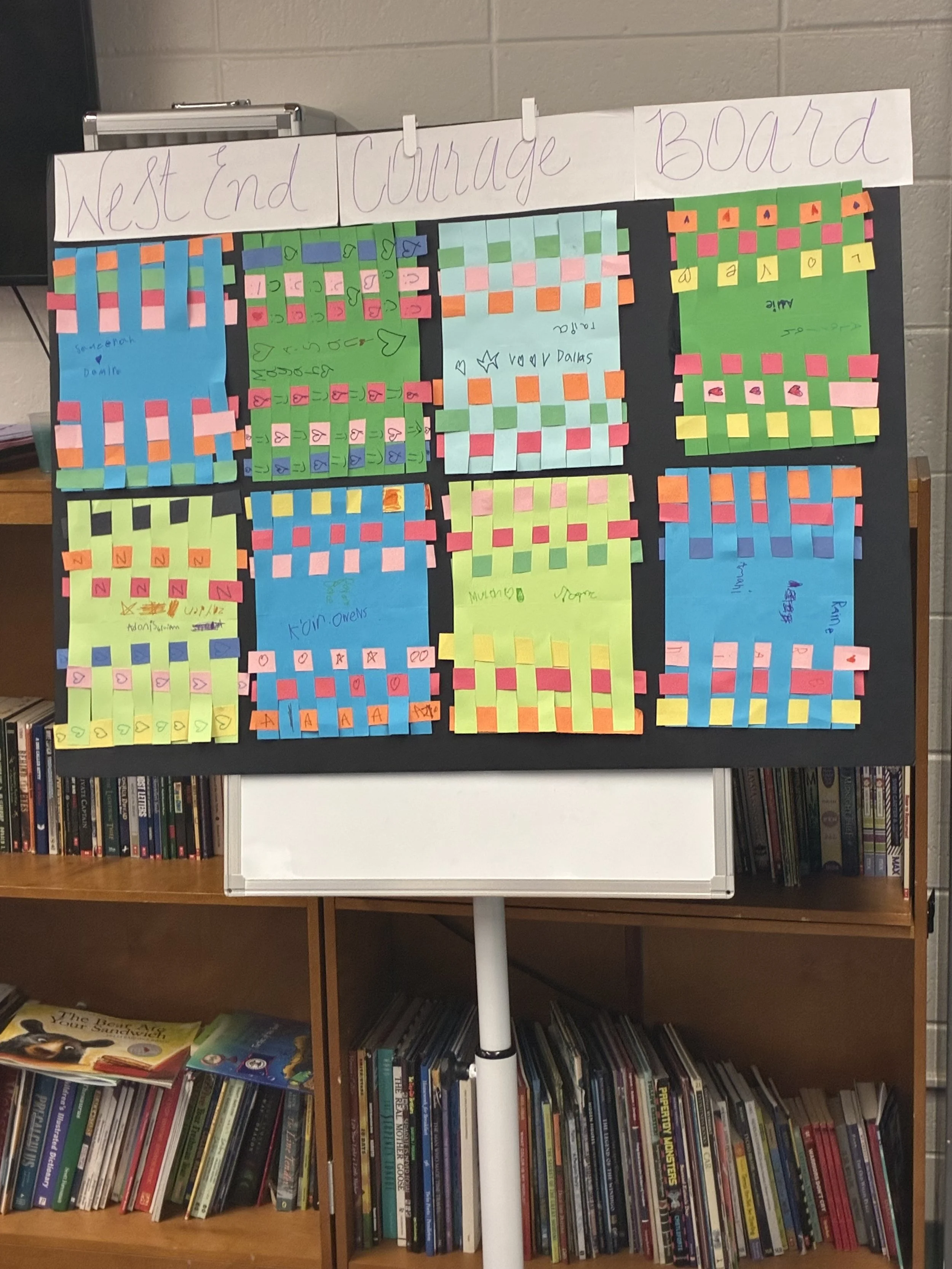A whiteboard with a black border clutch is displayed in front of a bookshelf. The whiteboard has several colored Post-it notes arranged into sections, with some handwritten titles including 'West End', 'Courage', and 'Board'. The bookshelf behind the