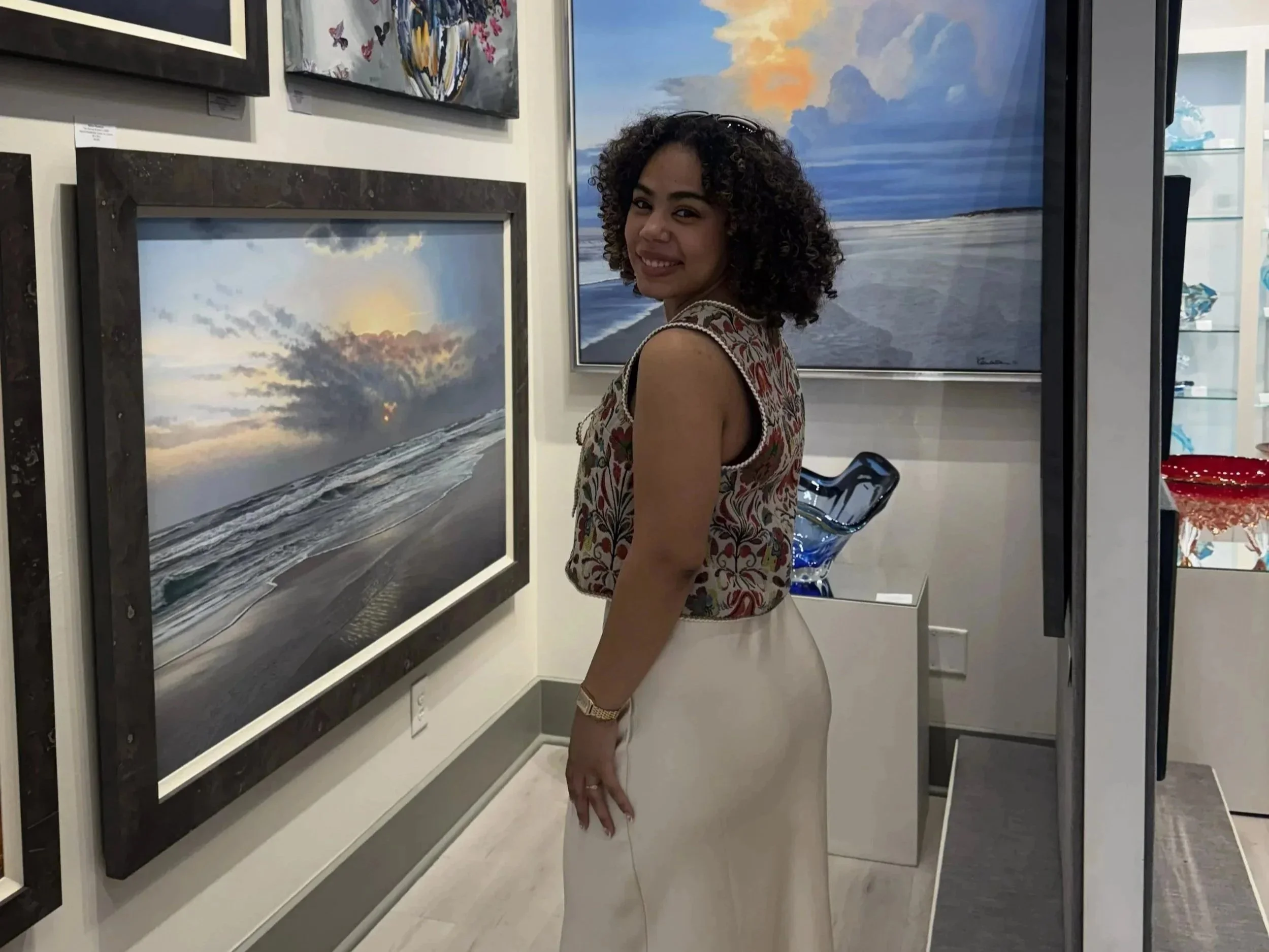 A woman with curly hair wearing a patterned sleeveless top, smiling at an art gallery, standing next to framed landscape paintings of the beach and sunset with clouds.