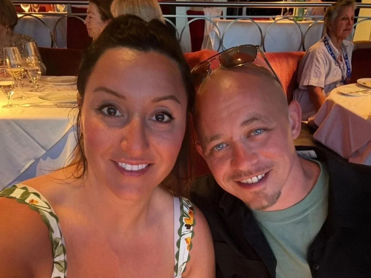 A woman and a man taking a selfie at a restaurant. The woman has dark hair and is smiling, wearing a white top with a floral pattern. The man is bald, wearing sunglasses on his head, and a black jacket over a gray shirt, also smiling.