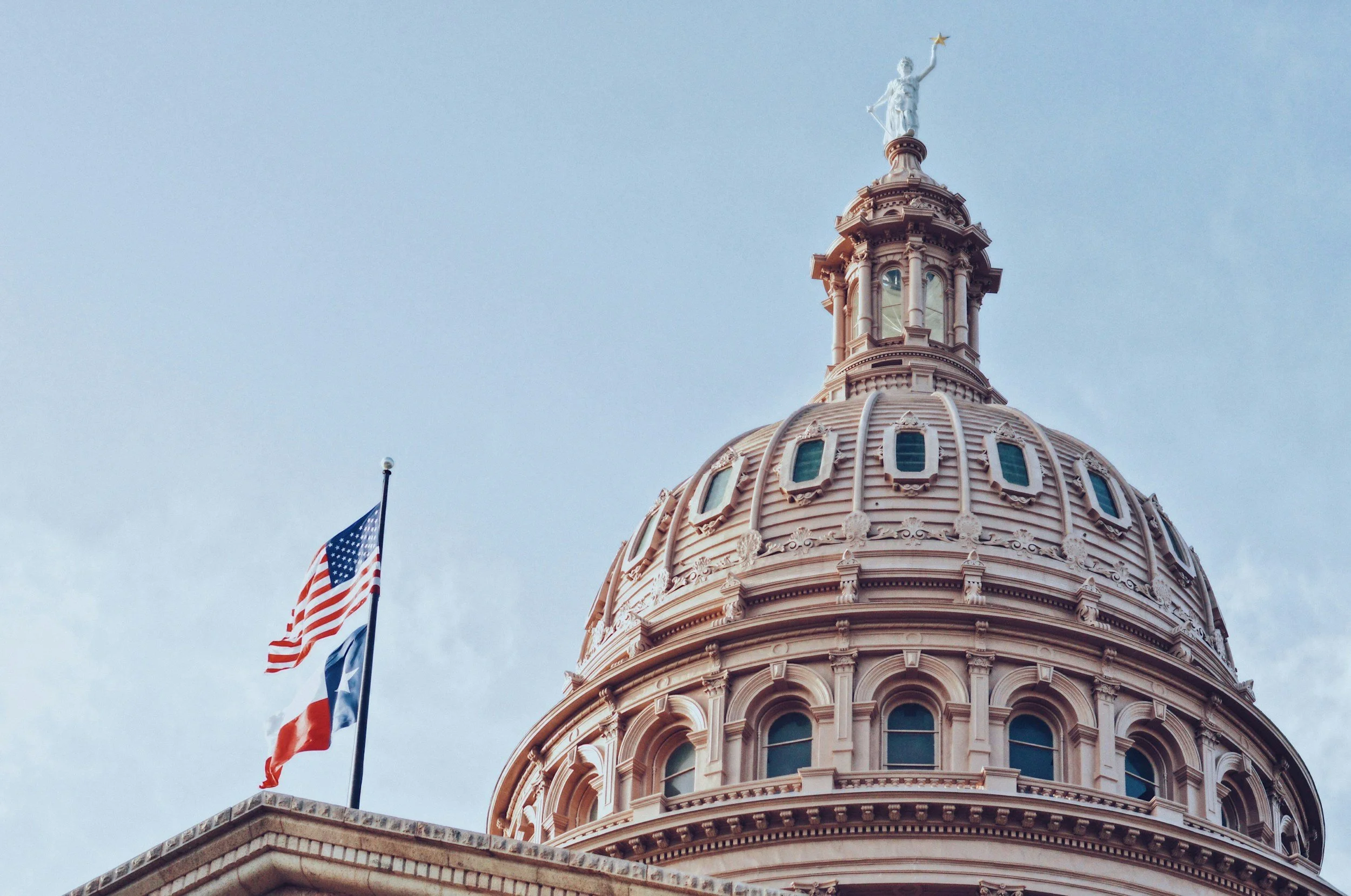 The image shows the dome of a capitol building with detailed architectural features, topped by a statue holding a star. American flags are visible next to the dome against a clear sky.