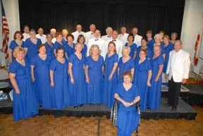 A large choir group of men and women dressed in blue gowns, standing on a stage, with two conductors or choir directors in white, posing for a group photo.