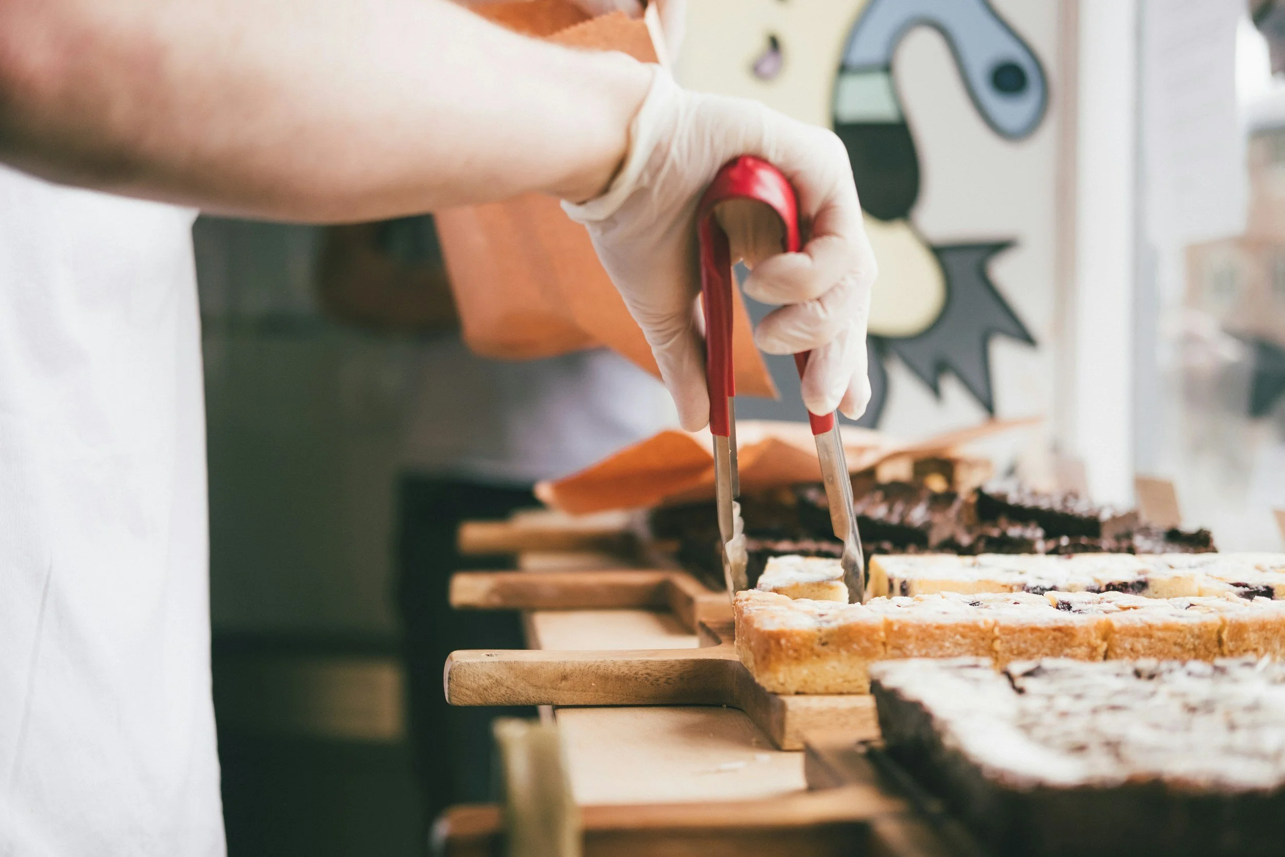 Person slicing layered dessert bars on a wooden table in a bakery or kitchen.