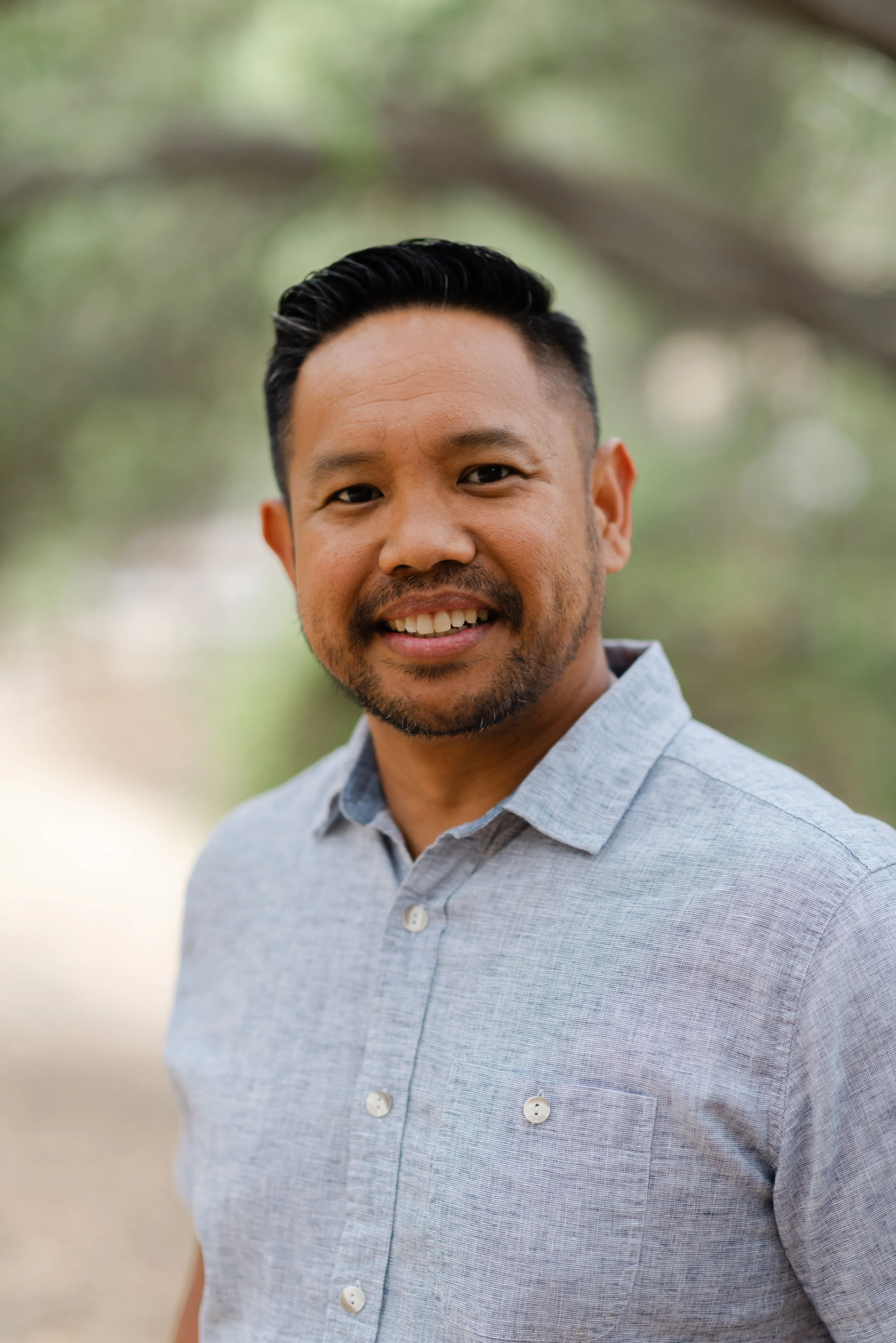 A man with short black hair, a trimmed beard, and a mustache smiling at the camera, wearing a light gray button-up shirt, standing outdoors with blurred green trees and foliage in the background.