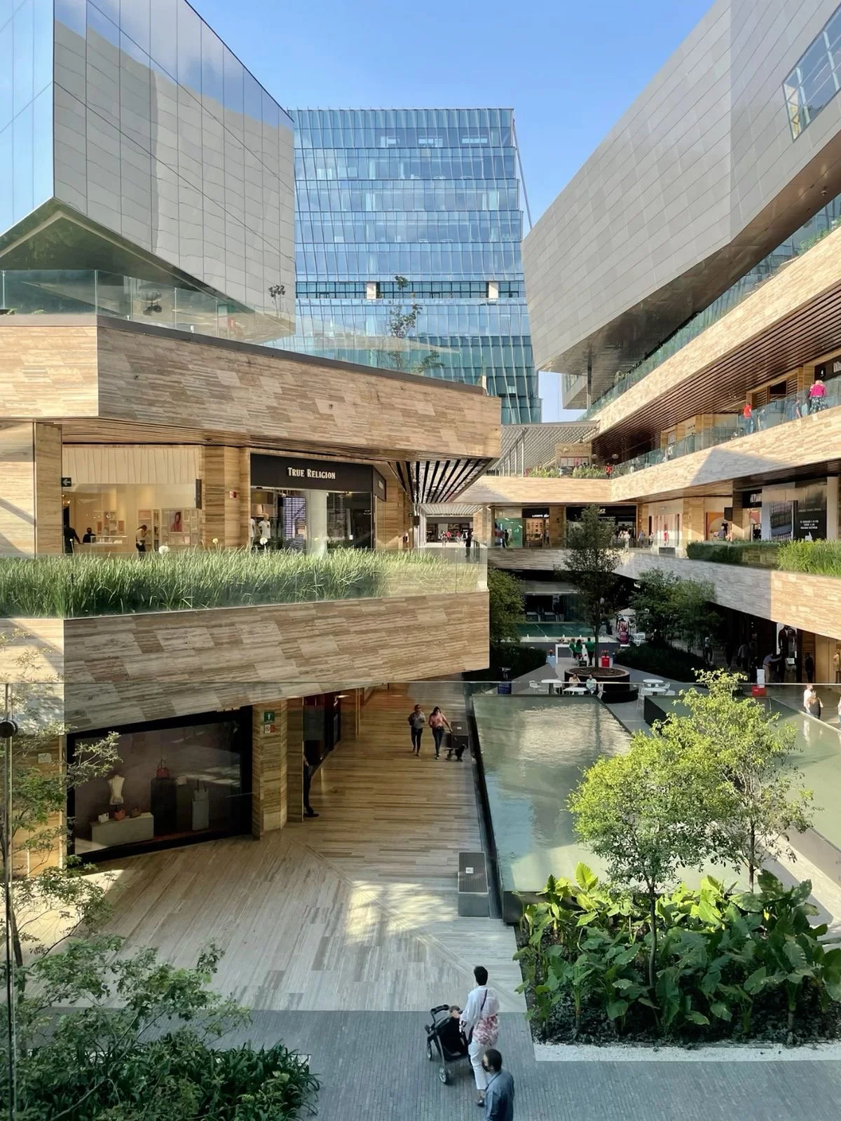 Modern multi-story shopping mall with glass and wood architecture, outdoor greenery, and open walkways with people shopping and walking.