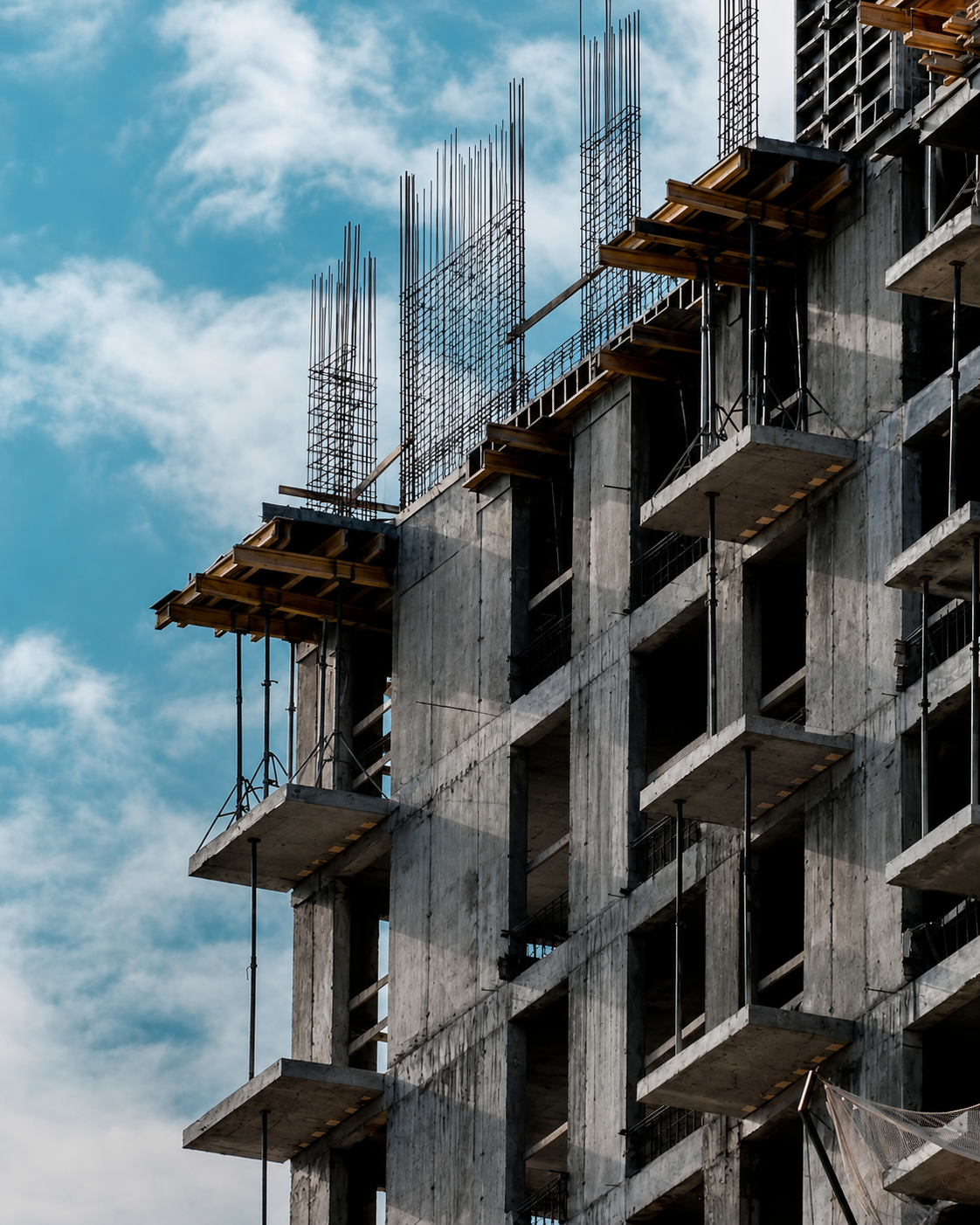 Under construction multi-story concrete building with exposed rebar and scaffolding against a partly cloudy sky.