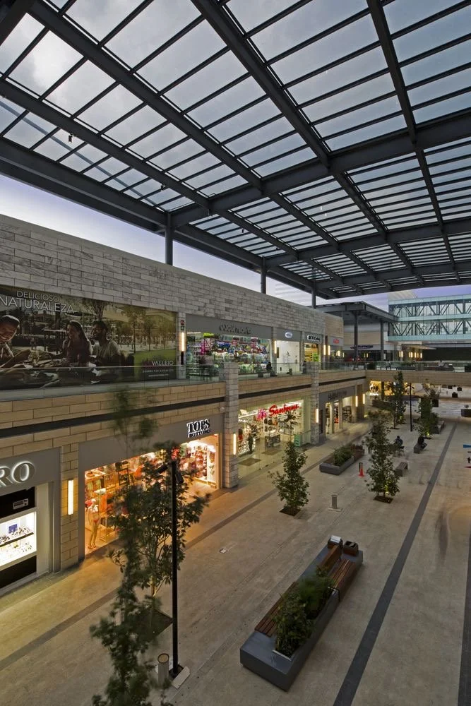 Indoor shopping mall with a glass roof, stores on a lower and upper level, benches, and potted trees along the walkway.
