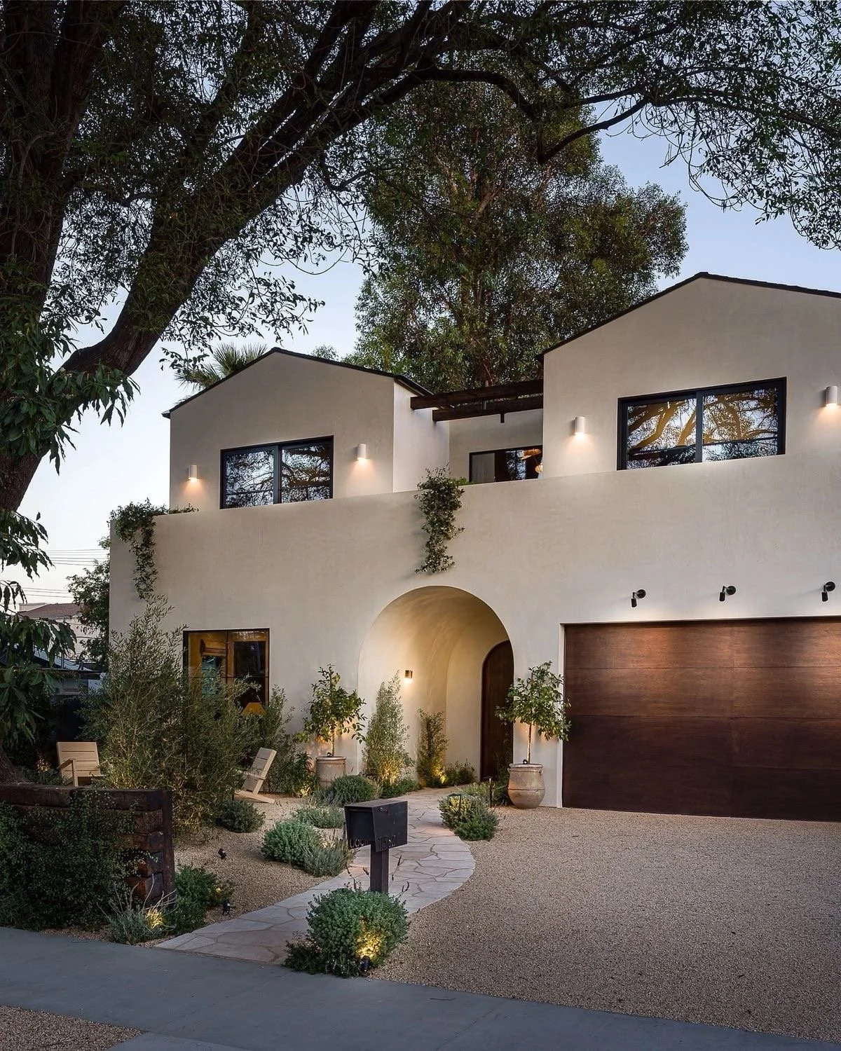 Modern two-story house with white stucco exterior, large black-framed windows, and a wooden garage door. The house is surrounded by landscaped garden with small trees, plants, and outdoor seating. Large trees are visible in the background, and exterior lighting illuminates the facade during dusk.