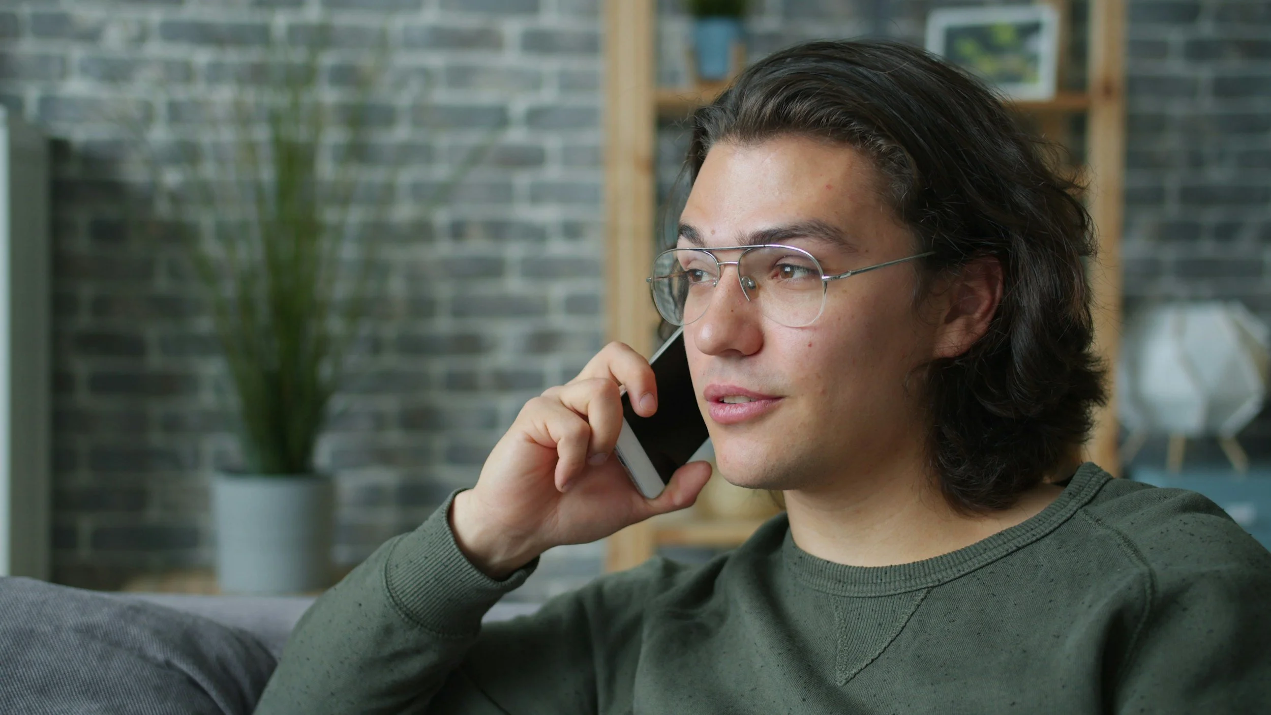 A young man with glasses and long dark hair talking on a cellphone indoors.